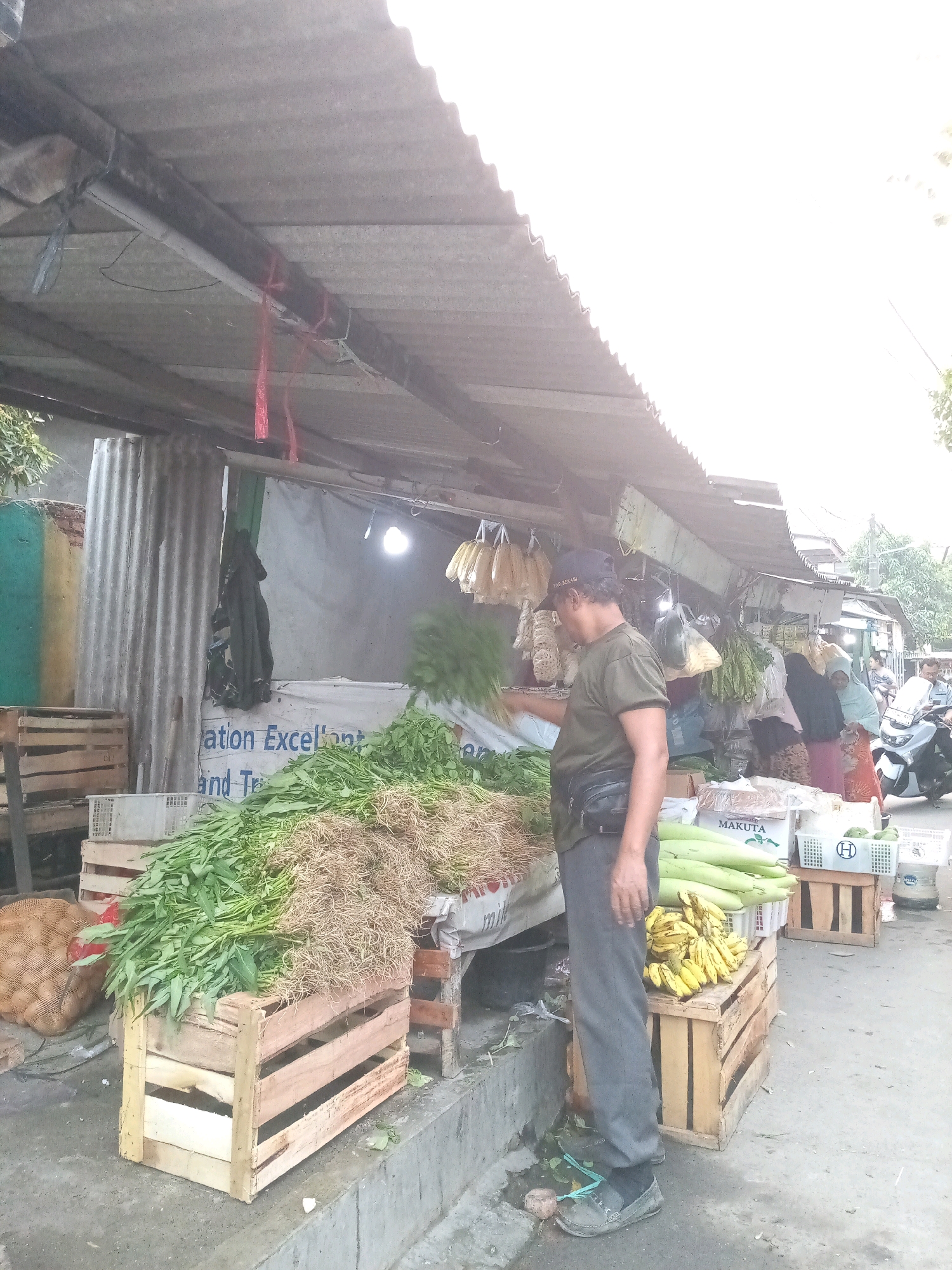 The image depicts an outdoor market stall in Bekasi, Indonesia. A man wearing a dark green t-shirt, grey pants, and a dark baseball cap stands behind a stall displaying various fresh produce. He is holding a bundle of green leafy vegetables. The stall has a corrugated metal roof supported by wooden beams. In front of him, there are several wooden crates filled with produce, including a large pile of green leafy vegetables with a mound of dry roots next to them, and a crate of yellow bananas. Behind the man, other market activities are visible, including people browsing and a motorcycle. The ground is paved with concrete, and there are some fallen leaves and debris. A white banner with text is visible behind the vegetables.
