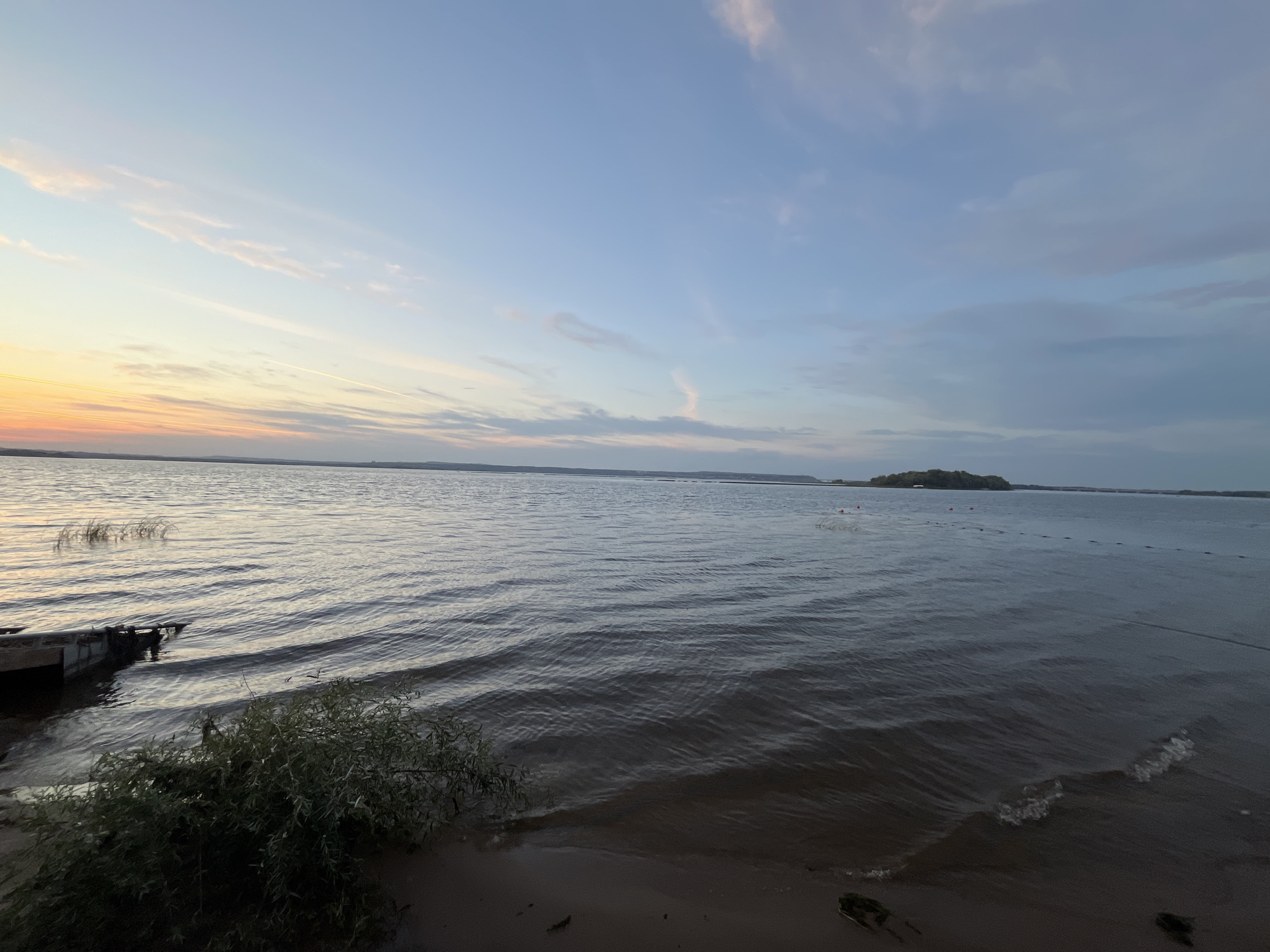 The image depicts a wide body of water, likely a lake or river, under a twilight sky. The sky transitions from warm hues of orange and yellow near the horizon on the left to a soft blue with scattered clouds on the right. The water is calm with gentle ripples, reflecting the sky's colors. In the foreground on the left, a section of a wooden dock is visible, with some vegetation growing near the shoreline. On the right side of the frame, the water extends to a distant horizon where land with trees is faintly discernible. Several buoys are scattered across the water in the mid-ground, indicating a marked area or perhaps a net. The overall atmosphere is serene and peaceful, characteristic of a sunset or sunrise over a natural landscape. The location is identified as the city of Shumeika, Russia.