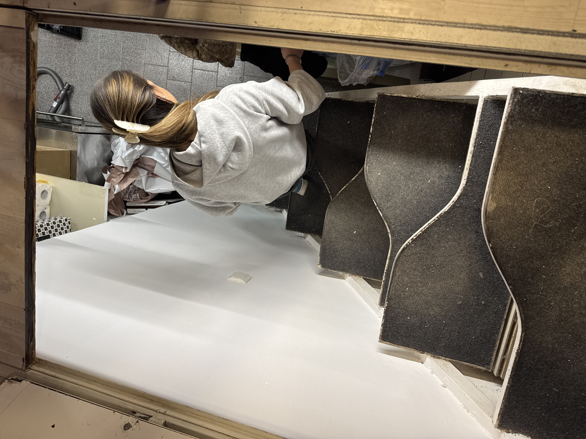 A woman in a grey hooded sweatshirt and hair clip stands next to a white countertop or platform with several dark grey-covered, possibly carpeted, shapes aligned along the right side. Behind her, in what appears to be a storage or service area, there's a vacuum cleaner, cardboard boxes, and other supplies. The view is captured from a high angle, looking down through a doorway or opening. The location is unknown, and there is no verifiable evidence linking this to Sofia, Bulgaria.
