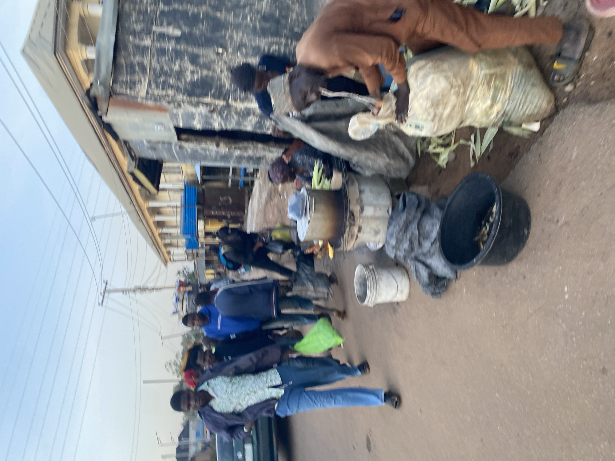 The image depicts a street scene in Gura Top, Nigeria, possibly a market area. In the foreground, a man in a brown traditional outfit is interacting with a large sack, appearing to be either filling it or retrieving something from it. Behind him, a woman is seated near cooking pots, suggesting food preparation. Several other individuals are visible in the background, some walking and others gathered around stalls or shops. The environment includes a dirt road, a building with a light-colored facade and columns, and makeshift structures made of corrugated metal and tarpaulin. The overall atmosphere suggests a bustling commercial or social gathering point.