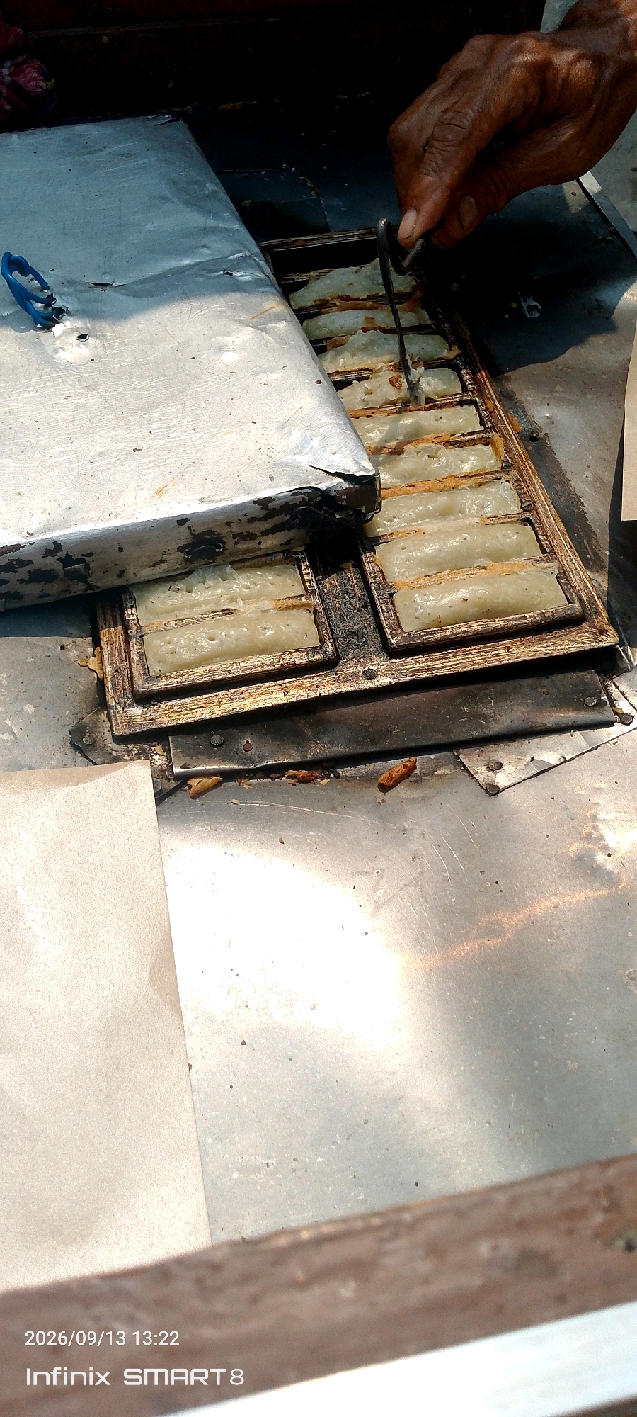 The image shows a close-up of a street food vendor in Serang, Indonesia, preparing traditional Indonesian snacks. The vendor is using a long metal tong to flip a series of small, rectangular pastries being cooked in a specialized mold. The pastries appear to be a light, creamy color, with some browning on the edges. The mold itself is made of a dark metal and has multiple rectangular slots, indicating it's designed for mass production of these snacks.
To the left of the mold, a large, somewhat dented metal cover is partially obscuring the cooking area. A blue plastic object, possibly a part of a tool or a clamp, is attached to the edge of this cover. The overall scene is set outdoors, as suggested by the bright sunlight casting shadows and illuminating the metallic surfaces. The ground surface is a rough, reflective metal, typical of a mobile food cart or stall. There are some crumbs and debris visible on the metal surface, indicating ongoing food preparation.
The timestamp "2026/09/13 13:22" and the text "Infinix SMART8" are overlaid on the bottom left of the image, indicating the date and time the photograph was taken, and the device used. The presence of the vendor's hand and the tongs actively engaged in cooking implies an active, operational food stall.