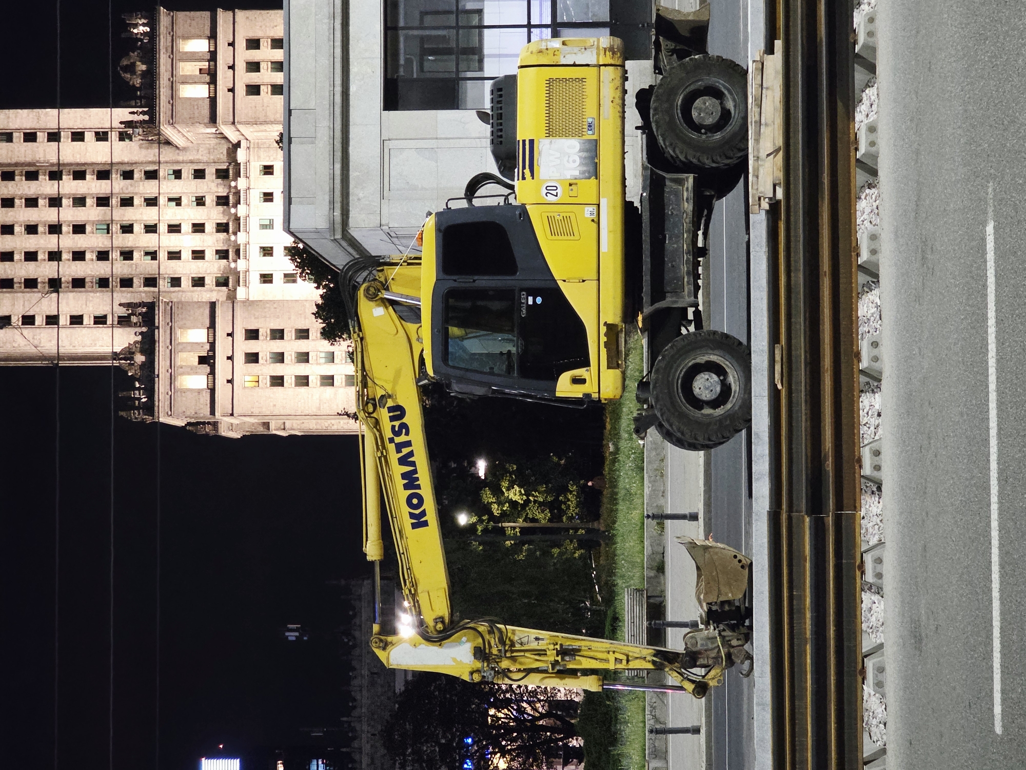 An outdoor, nighttime, eye-level, full shot captures a yellow Komatsu excavator parked on a paved surface next to trolley tracks in Warsaw, Poland. The excavator is oriented horizontally, facing right. Its long yellow arm is extended forward. The machine's cabin is dark, with a "GALEO" logo visible. The side of the excavator displays "20 PW 160". Behind the excavator, a large, light-colored building with many windows is illuminated. Trees and streetlights are visible in the background, along with the faint outline of another building on the left. The foreground shows the asphalt road and trolley tracks. There are no people visible in the frame.