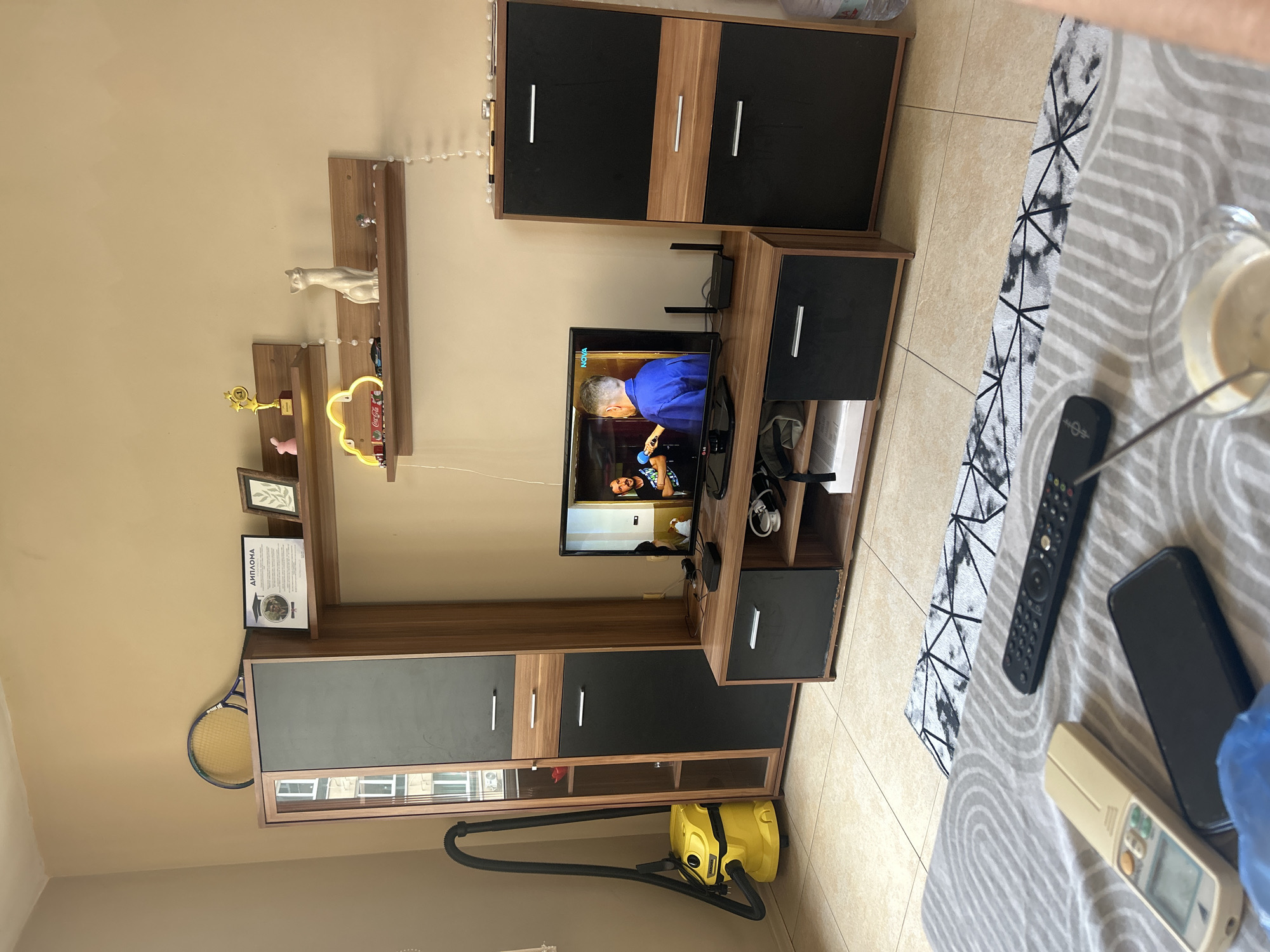 The image depicts a living room scene, likely in Plovdiv, Bulgaria, featuring a modern entertainment unit against a beige wall. A large, dark-fronted cabinet with wooden accents stands to the left, housing a yellow Karcher vacuum cleaner at its base. To the right of this cabinet is a tall, dark-fronted unit with drawers and cabinets. A flat-screen television rests on a low-lying entertainment console between these cabinets, displaying two men, one holding a microphone. Above the television, wooden shelves are mounted on the wall, adorned with various decorative items including a tennis racket, a diploma, a small trophy, a pink bunny figurine, a cloud-shaped neon light, a white cat figurine, and a miniature car. A bedside table is partially visible in the foreground, with a remote control, a smartphone, and a glass of coffee on its surface. The flooring consists of beige tiles.