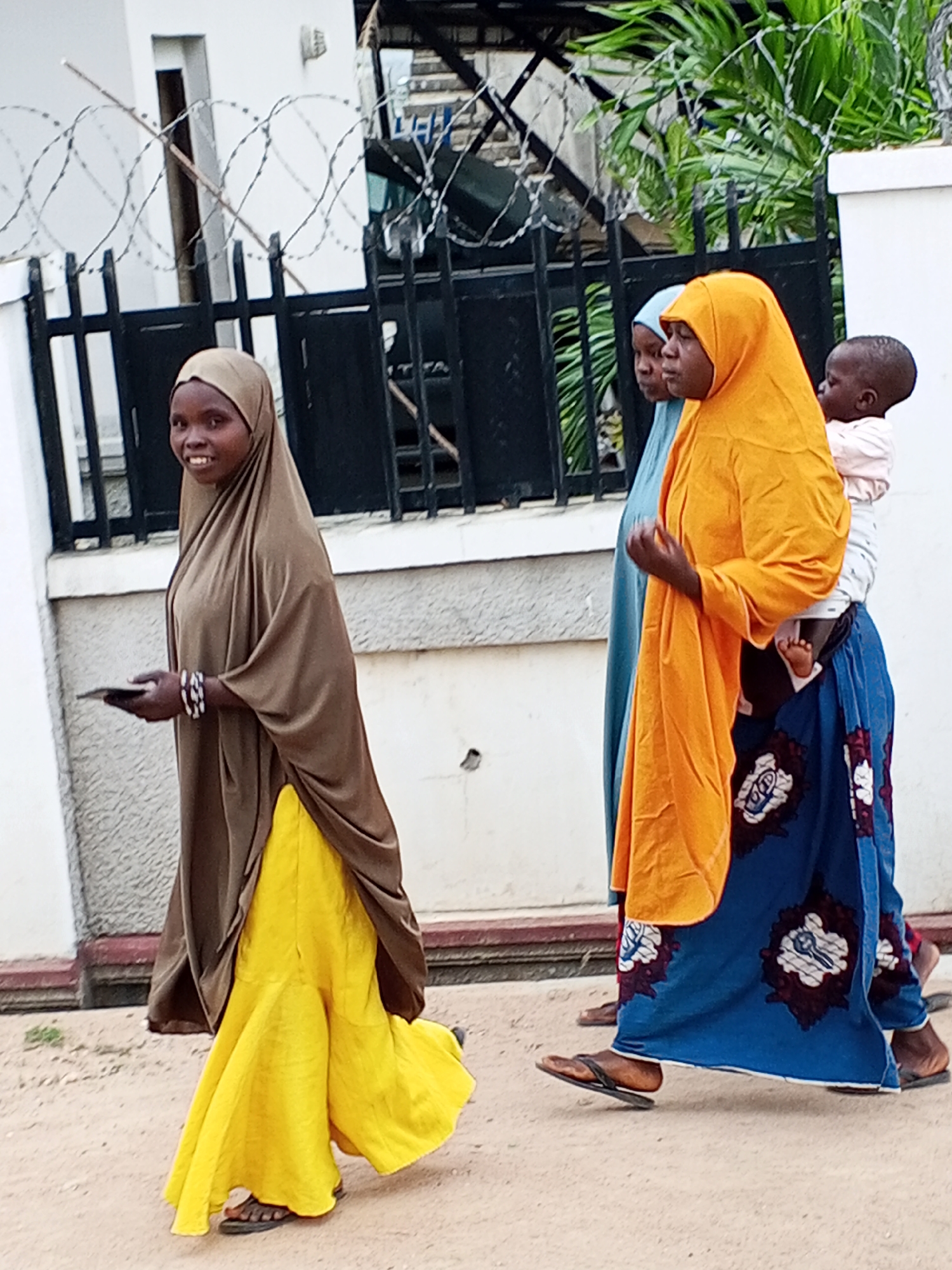 Three women walk along a dirt road in Adonkolo, Nigeria. The woman in the foreground wears a brown headscarf and a yellow dress, and holds a black object. Behind her, a woman in a blue headscarf follows. The third woman, carrying a baby, wears an orange headscarf and a blue patterned skirt. A fence with barbed wire and a building are visible in the background.