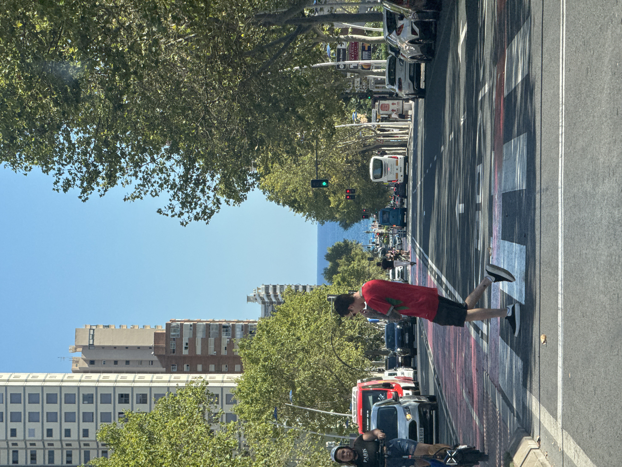 This image captures a street scene in Benidorm, Spain, likely along the main coastal avenue given the tall buildings and proximity to the sea. In the foreground, a pedestrian crossing with red and white markings is prominent. A man in a red t-shirt and black shorts is walking across the pedestrian crossing, looking down and to his right. To his left, a woman wearing a black t-shirt with "PARIS" on it and denim shorts is standing on a personal electric transporter, leaning on a white car. A red fire truck is parked behind the white car. In the background, a tree-lined avenue stretches towards the sea, with several vehicles including a white bus. The sky is clear blue. Tall buildings, characteristic of Benidorm's skyline, flank the street on the left. The overall scene depicts a sunny day in a bustling urban environment near the coast.