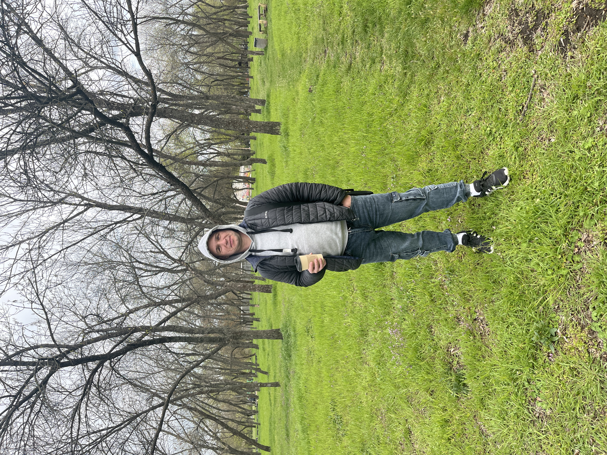 A young man stands in an outdoor park setting, looking directly at the camera with a slight smile. He has short, dark hair and light stubble. He is dressed in a light grey hooded sweatshirt, a black quilted puffer jacket with a visible "HI-TEC" logo, blue jeans, and black and white athletic shoes (the brand "NIKE" is discernible on the sole and side). His left hand is in his pocket, and he holds a light brown paper cup or container in his right hand.
The scene features a wide expanse of bright green grass, indicating early spring or late winter. In the background, a long row of tall, deciduous trees with bare branches lines what appears to be a path or boundary. The sky is overcast and bright, providing soft, diffused lighting. Beyond the trees, some faint structures, possibly park amenities like benches, are visible in the distance. The overall environment suggests a tranquil, open-air location within Kryvyi Rih, Ukraine.