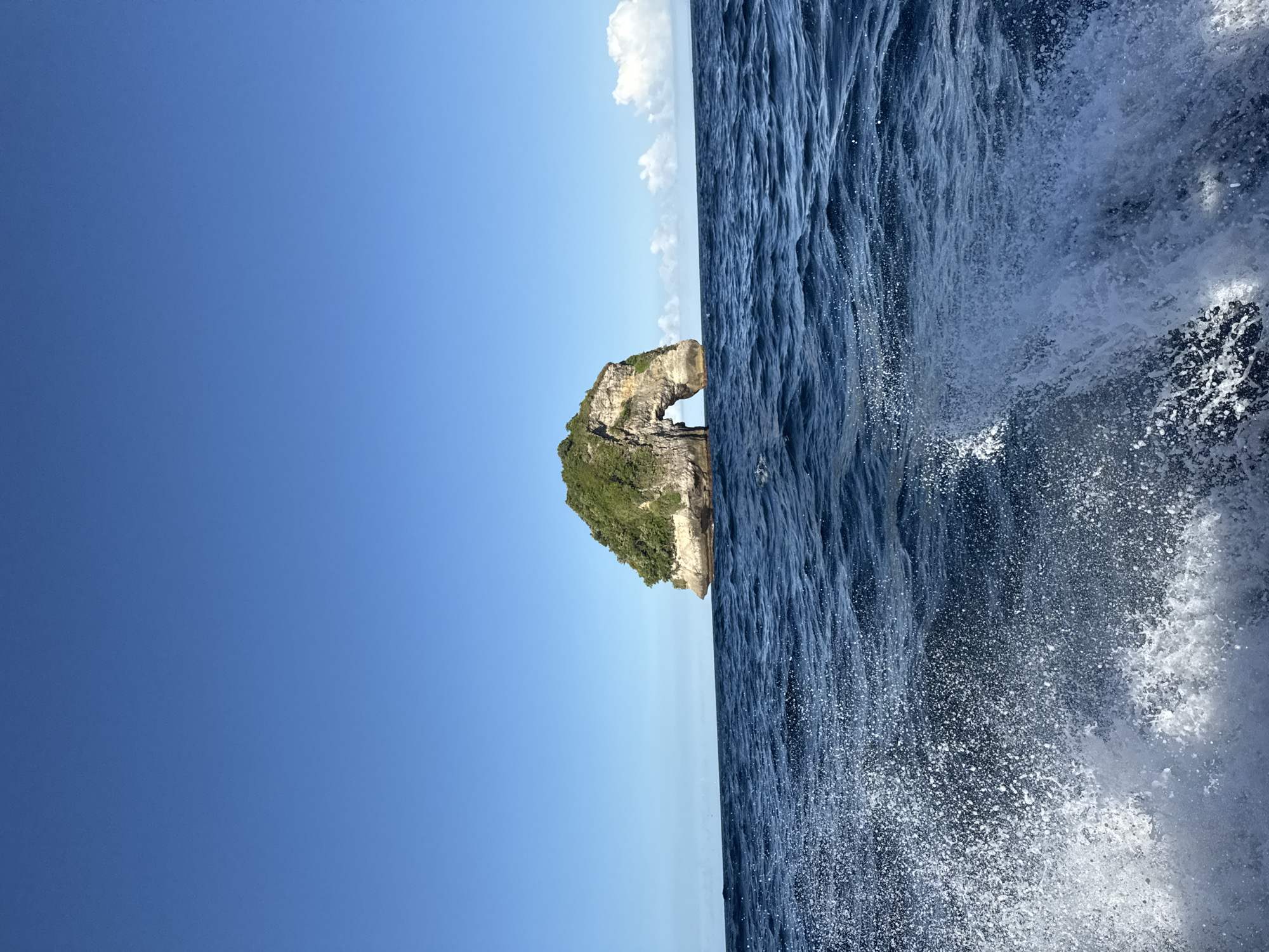 The image shows a rock formation with a natural arch in Nusa Penida, Indonesia. Lush green vegetation covers the top of the rock. The sky is a clear blue with a few scattered white clouds near the horizon line. The ocean is dark blue and rough, with splashes of white water visible on the right side of the image, suggesting waves crashing. The image appears to be taken from a moving boat, with the ocean spray coming towards the camera.