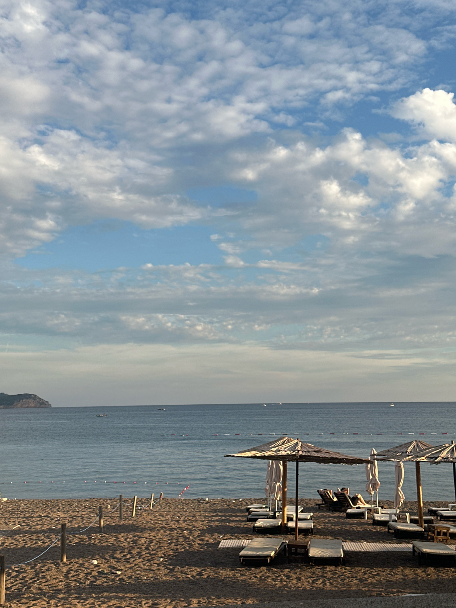 The image captures a serene beach scene in Boreti, Montenegro, under a sky filled with scattered clouds. In the foreground, a sandy beach is lined with rows of empty lounge chairs and thatched umbrellas, suggesting a tranquil atmosphere. A few figures are visible reclining on lounge chairs under the umbrellas, appearing relaxed. The sea stretches out to the horizon, calm and blue, with a few distant boats dotting the water. To the left, a hilly landmass juts into the sea, providing a scenic backdrop. The overall impression is one of a peaceful coastal resort, likely during late afternoon or early evening given the lighting.