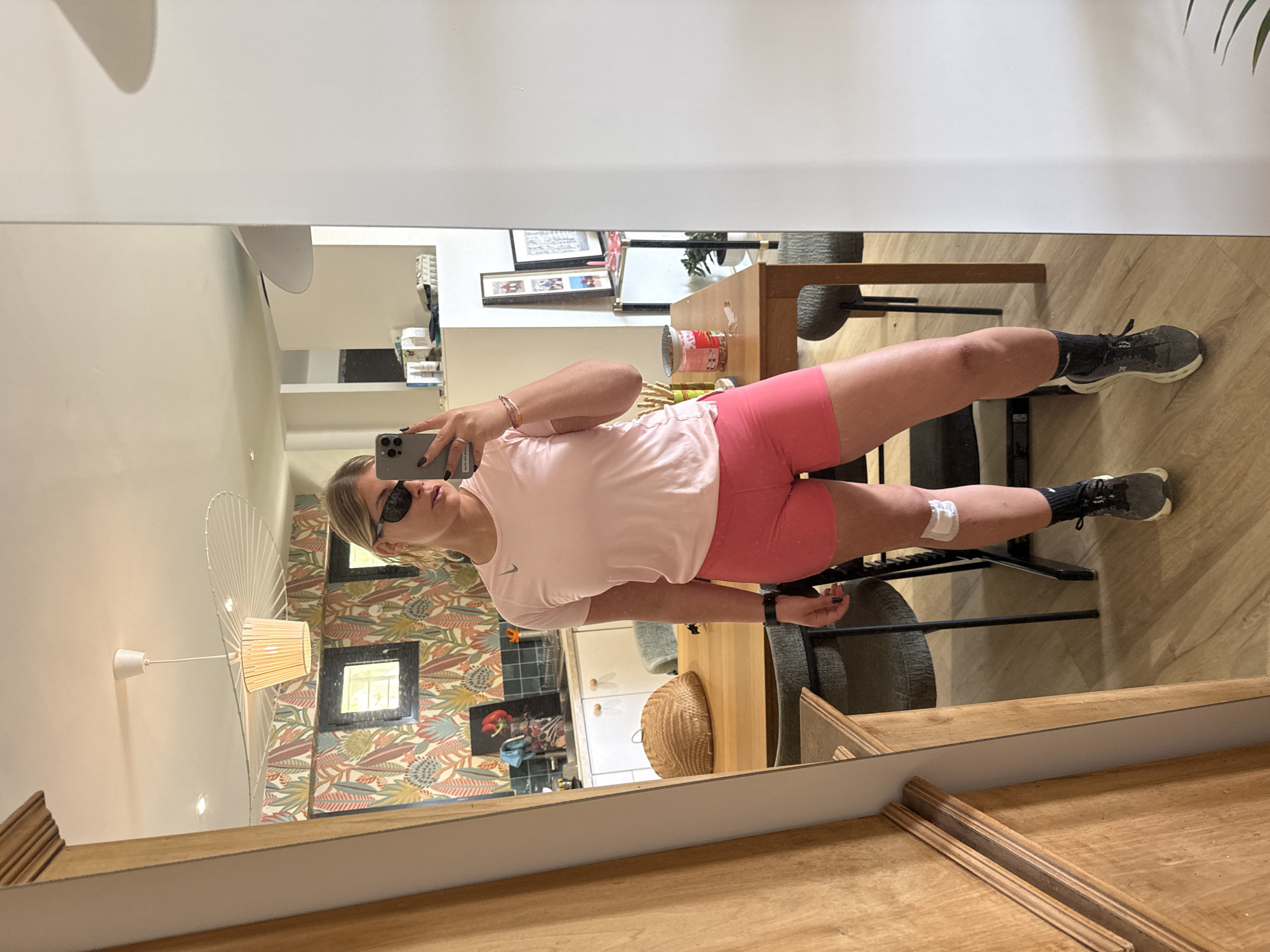 The image shows a woman in a light pink Nike t-shirt and pink shorts taking a selfie in a mirror. She is also wearing black athletic shoes, black socks, sunglasses, and has a bandage on her right calf. The reflection in the mirror reveals a portion of the room behind her, including a wooden dining table with chairs, a kitchen area with white cabinets and colorful wallpaper, a ceiling light fixture, and framed pictures on a shelf. The reflection suggests an indoor setting. While a specific location in Rousset, France cannot be determined from the image, the presence of typical French indoor elements (e.g., style of furniture) suggests that the photo was taken indoors in that country.