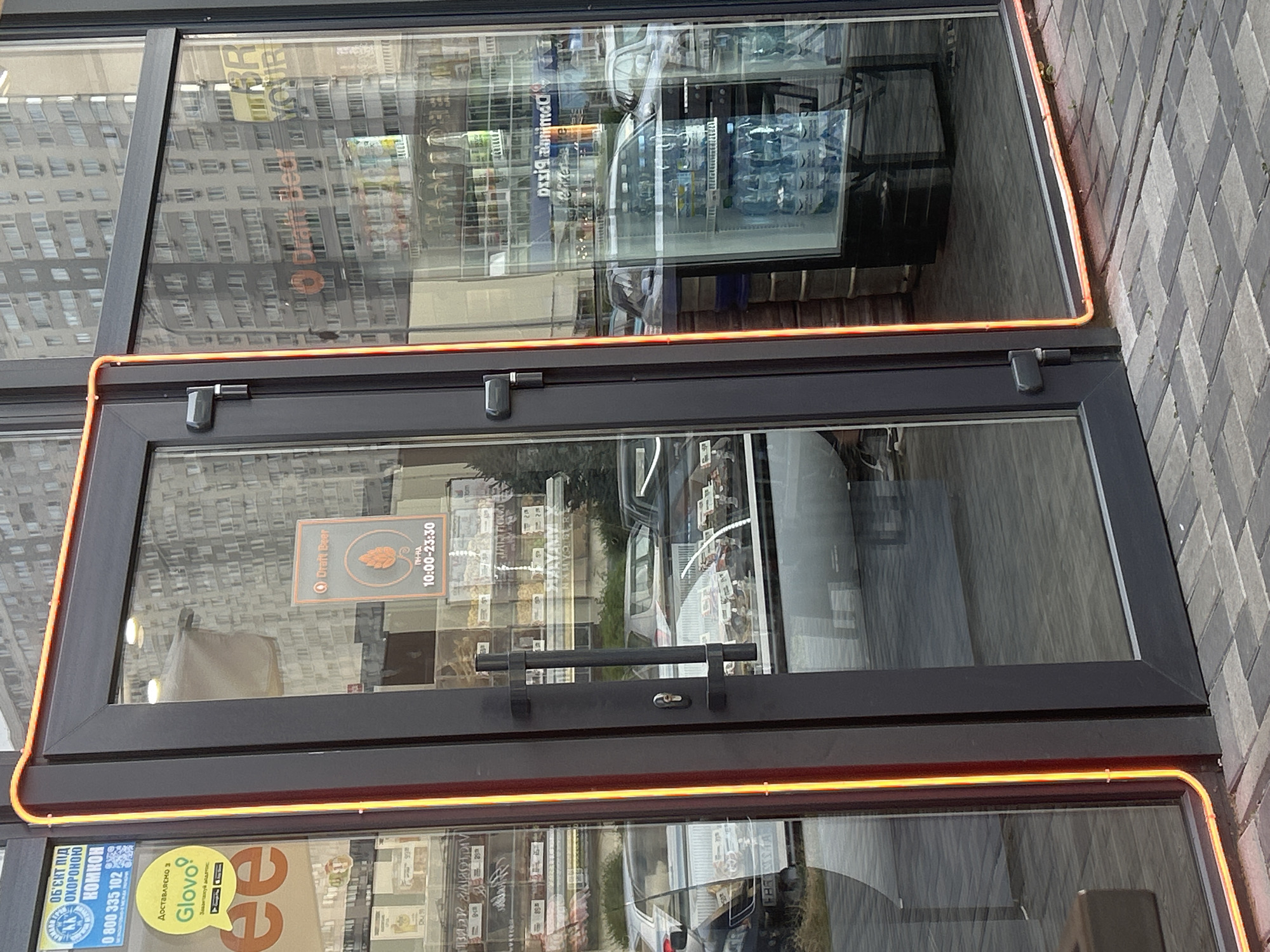 The image shows the entrance to a store in Brovary, Ukraine. The entrance features a double glass door with black frames. A neon orange light runs along the edge of the door frame. Inside the store, shelves are visible stocked with various items, and a refrigerated display case is filled with bottled water. Reflections in the glass show buildings and cars outside. A sign on the door indicates "Draft Beer" with operating hours from 10:00 to 23:30. Another sticker advertises delivery through Glovo. The ground outside is paved with interlocking bricks.