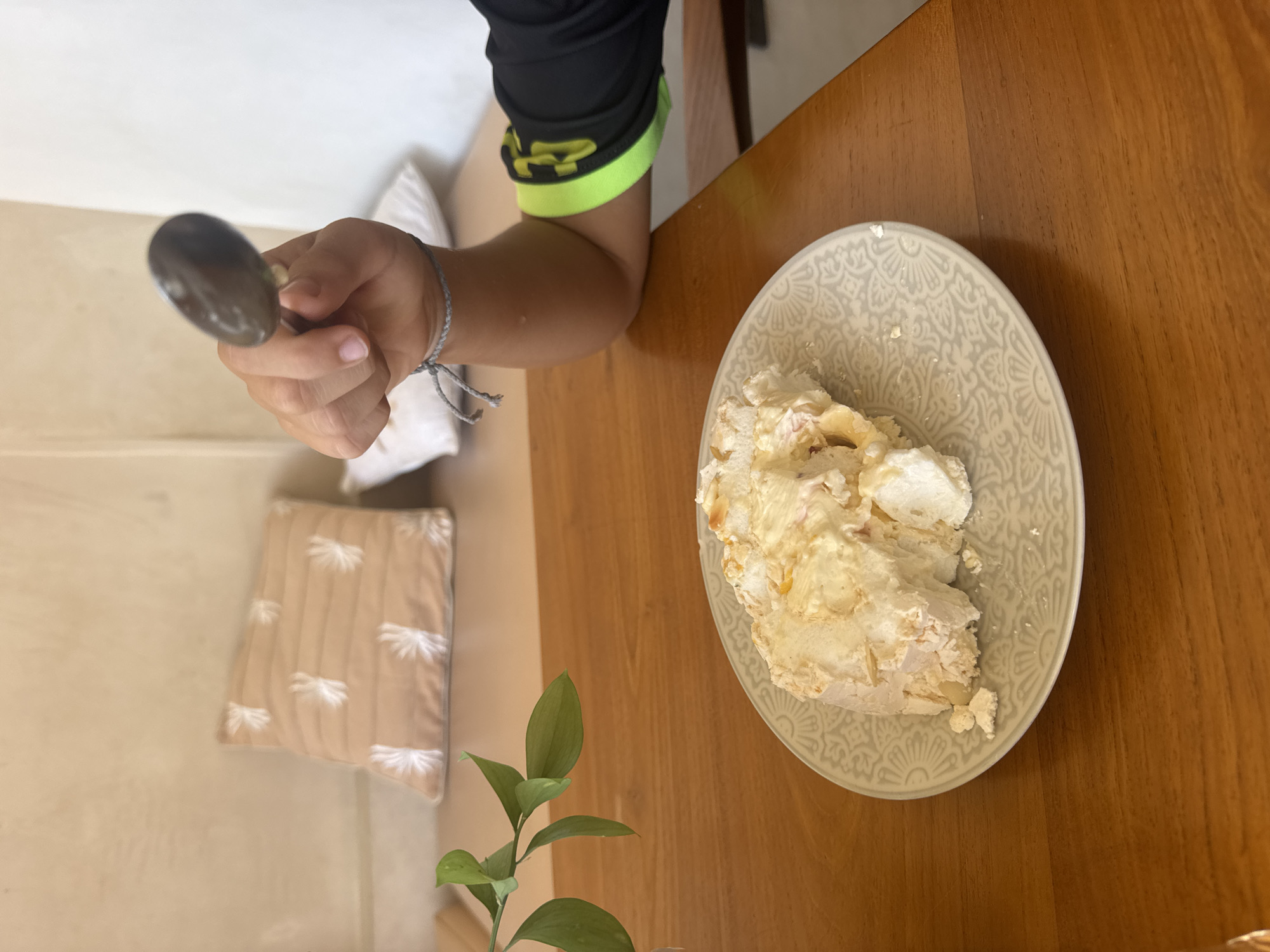 An overhead shot shows a slice of meringue cake on a patterned grey plate, resting on a wooden table. To the left, a sprig of green leaves extends into the frame. In the background, a light brown pillow with white floral embroidery is visible on a built-in bench. A person's arm is in the right side of the frame, holding a spoon with a dark, indistinct object on it, poised above the cake. The person wears a black long-sleeved shirt with a bright neon green band around the cuff and a grey string bracelet. The scene appears to be indoors, with soft, natural light illuminating the space. The setting suggests a cafe or restaurant in Kuta Selatan, Indonesia.