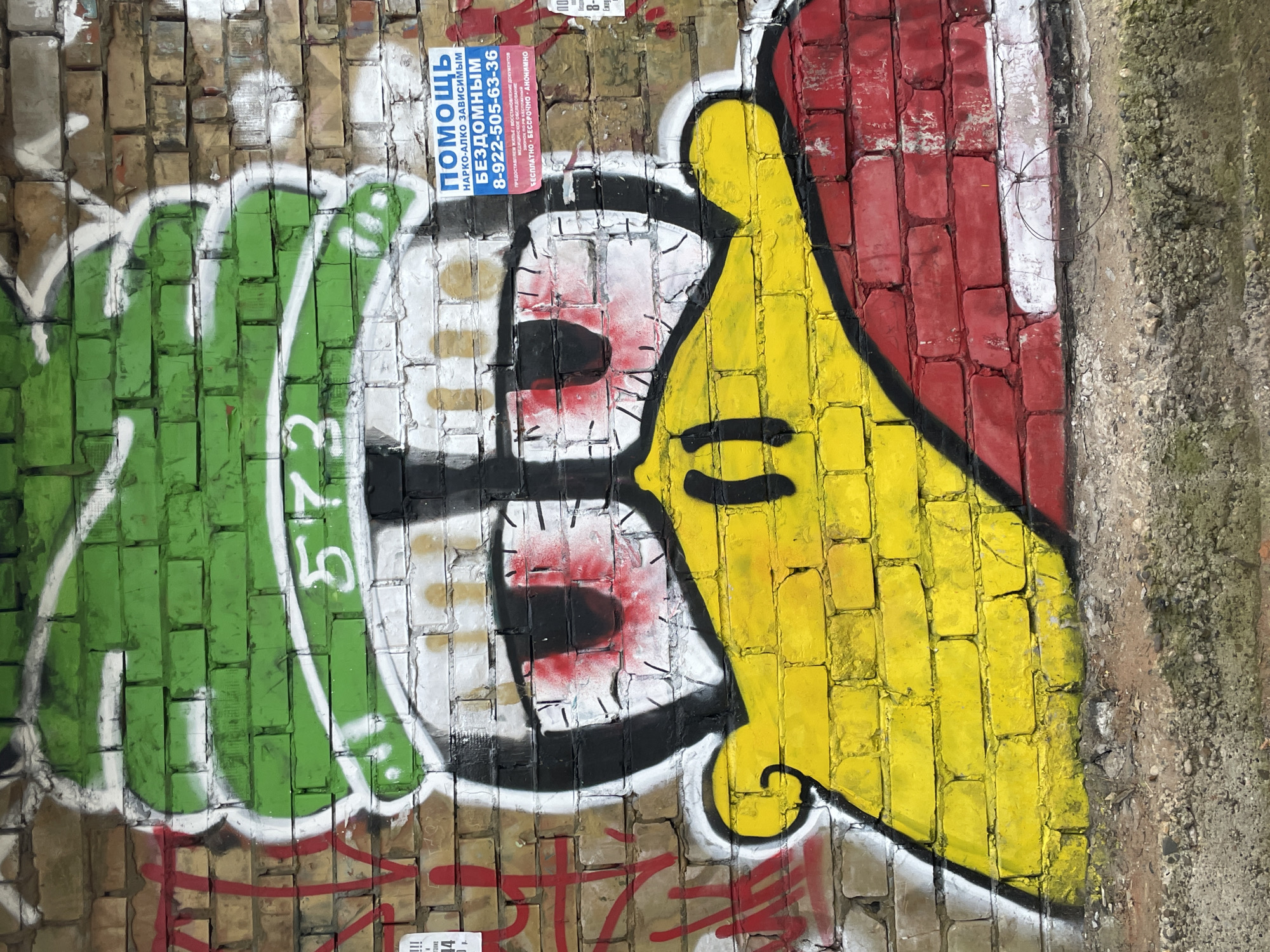 This image displays a vibrant graffiti artwork on a worn brick wall in Izhevsk, Russia, captured during daytime. The central subject of the graffiti is a cartoonish character, possibly a bird or a creature, depicted with a bright green left side, a white face featuring large, red-tinged eyes with black pupils, and a yellow right side that suggests a beak or horn. The entire character is outlined boldly in black. The number "573" is visible in white paint on the green portion of the graffiti.
Above the artwork, a blue flyer or poster is partially adhered to the brick wall. This flyer provides information about social assistance, prominently featuring the word "ПОМОЩЬ" (HELP). The text further specifies services for "НАРКО-АЛКО ЗАВИСИМЫМ" (DRUG-ALCOHOL ADDICTS) and "БЕЗДОМНЫМ" (HOMELESS), including offerings like housing, document restoration, and medical examination. A contact phone number, "8-922-505-63-36," is clearly printed, alongside assurances that the services are "БЕСПЛАТНО" (FREE), "БЕССРОЧНО" (INDEFINITE), and "АНОНИМНО" (ANONYMOUS). Other faint numbers, "14" and "10," are also visible elsewhere on the wall.
The brick wall itself consists of light brown bricks, many of which are covered by paint or show signs of wear, dirt, and weathering, characteristic of an urban environment. The combination of the expressive graffiti, with its potentially melancholy eyes, and the flyer advertising help for vulnerable individuals creates a poignant scene that highlights societal challenges within an urban setting.