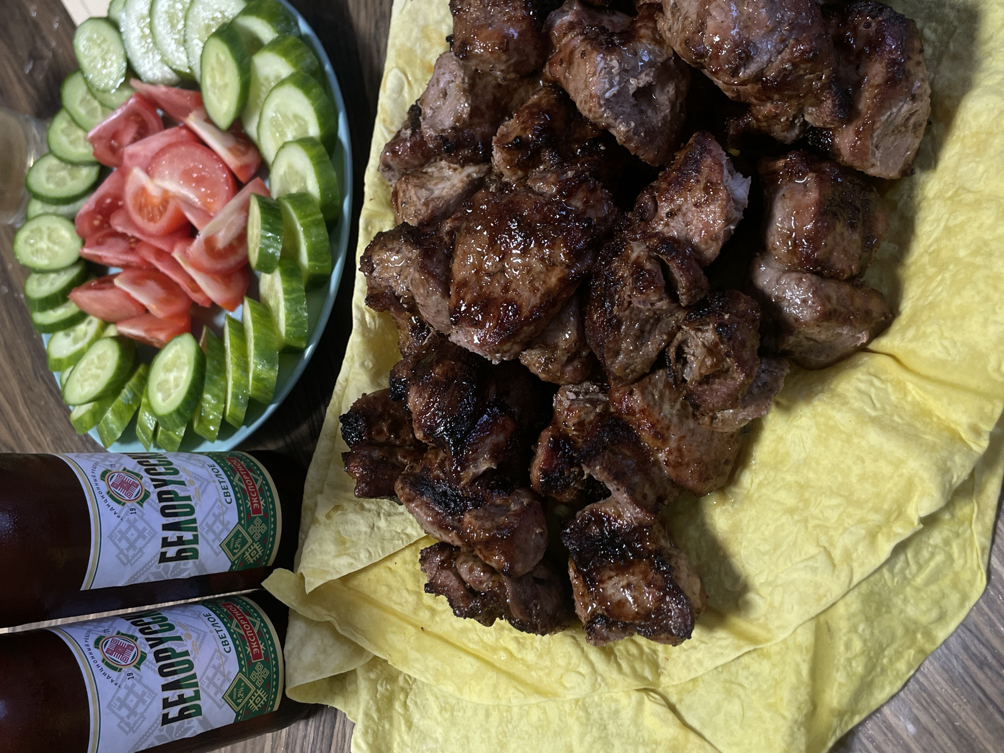 The image shows a meal consisting of grilled meat, sliced cucumbers and tomatoes, and two bottles of beer. The meat is piled on a yellow flatbread. The plate of cucumbers and tomatoes is arranged with slices alternating between the two vegetables. The beer bottles have labels in Russian. The location is Kuyeda, Russia.