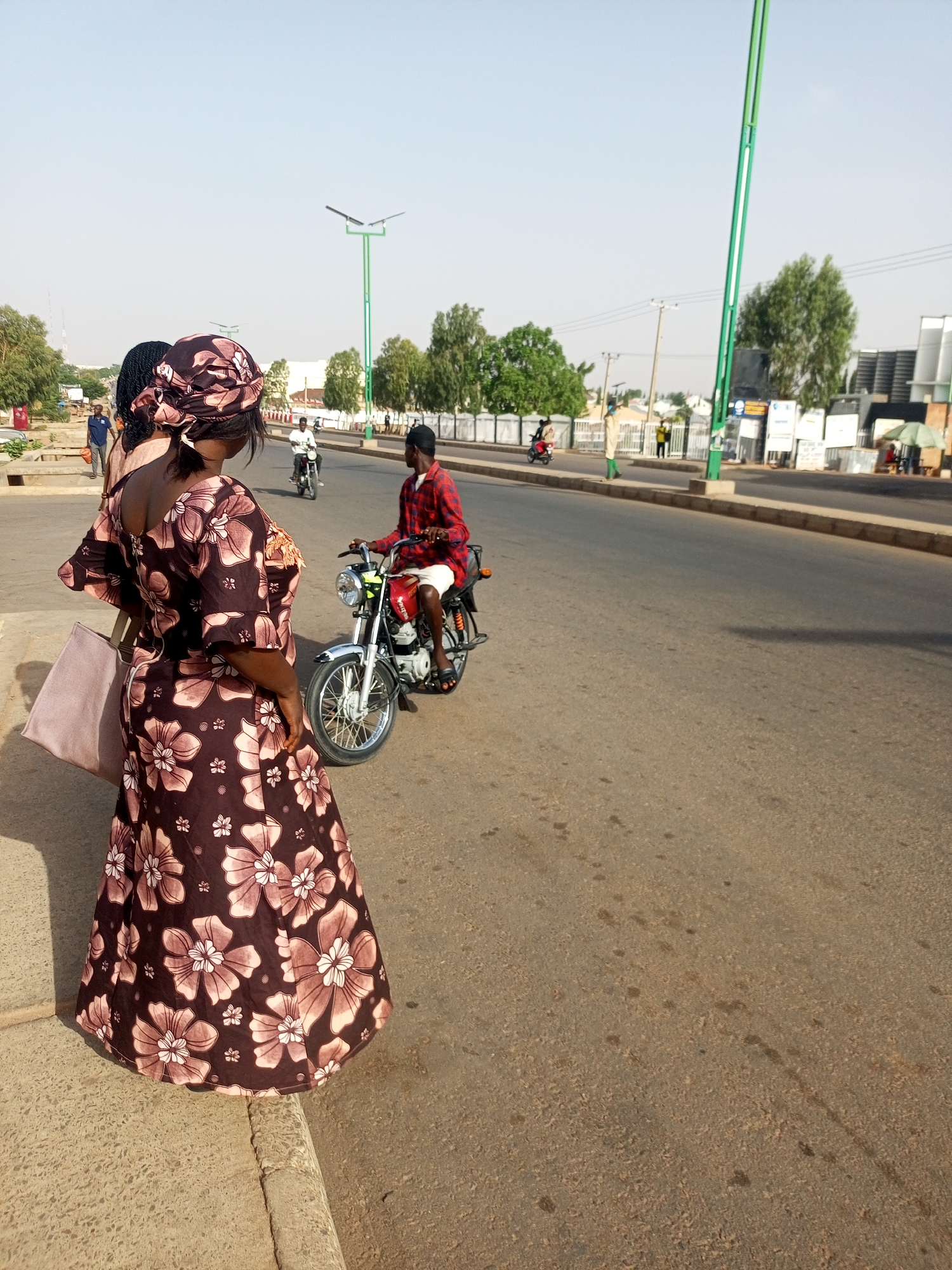 The image is taken in Gombe, Nigeria, and shows a woman with a brown floral print dress and head covering standing on the side of a road. She is holding a large, light-colored purse on her left shoulder. The road is asphalt and has a curb on the left side where the woman is standing.
In the background, a man in a red checkered shirt and black baseball cap rides a red motorcycle down the road. There is another motorcycle further down the road, as well as other people. There are tall green lampposts along the side of the road, and various buildings and trees in the distance. The sky is clear and light blue. The scene is bathed in sunlight.