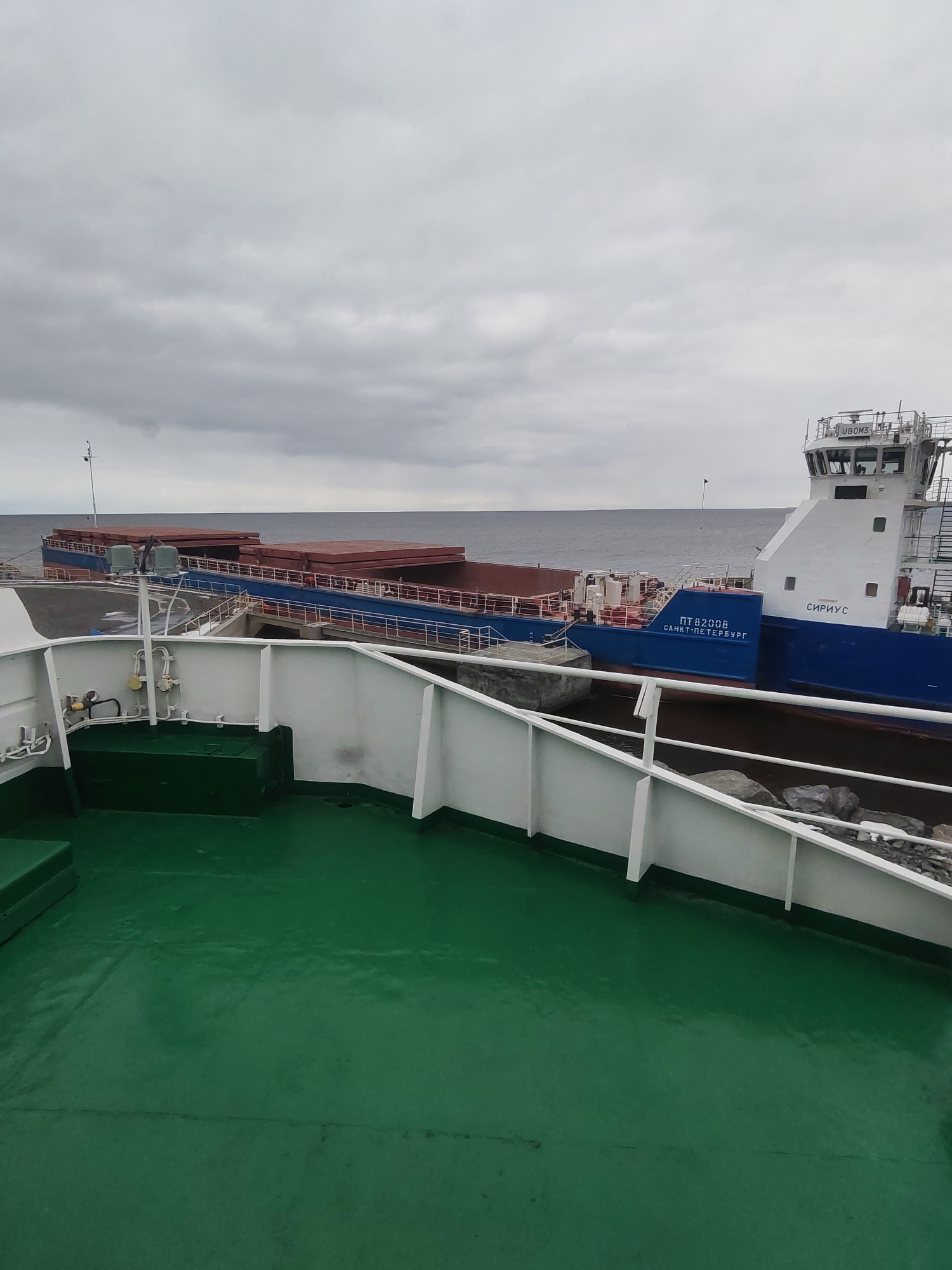 This image captures a view from the deck of one ship towards another ship moored or docked, with open water and a cloudy sky in the background. The scene appears to be at a port or a coastal area, likely in Shcheleiki, Russia, given the context.
In the foreground, the deck of a vessel is visible, characterized by its vibrant green paint and white railings. Several pipes and fixtures are mounted along the interior of the white railing structure.
The main subject is a blue and white cargo ship in the middle ground, viewed from its port side. This ship has large, reddish-brown cargo hold covers on its deck. Its superstructure, featuring a navigation bridge, is white with visible windows and equipment. The ship's name, "СИРИУС" (Sirius), is clearly painted in white letters on the blue hull near the stern. Below the bridge, the registration number "ПТ82008" and the homeport "САНКТ-ПЕТЕРБУРГ" (Saint Petersburg) are also visible on the blue hull. On the top of the bridge, the alphanumeric "UBOM3" can be seen.
Beyond the ships, the dark, calm waters of what appears to be a sea or large lake extend to the horizon. The sky is entirely overcast with grey clouds, indicating a gloomy or typical cloudy day. The area adjacent to the moored ship shows some rocky terrain, suggesting a natural shoreline or a breakwater. No people are visible in the frame. The overall atmosphere is calm and industrial, highlighting marine transport.