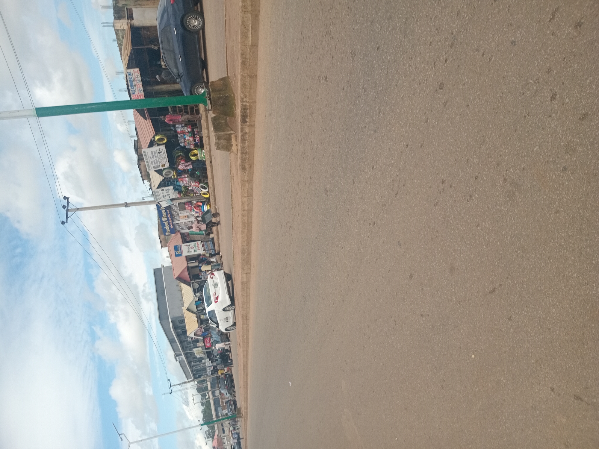 This is a photograph taken from a street level perspective, showing a busy market area in Bukuru, Nigeria. The foreground is dominated by an asphalt road with a concrete curb and raised concrete barriers separating the road from the sidewalk. In the middle ground, a white sedan is parked facing away from the camera. Beyond the sedan, a row of market stalls and shops are visible. These structures have various roofing materials, including corrugated metal and what appear to be tiled roofs. Signs for businesses such as "Prudent Wines", "LG", and "R.L.C CHUDI INT." are visible. Numerous people are present, some standing in front of the shops, others appearing to be engaged in transactions or walking. Objects like tires, fruit juice bottles, and other merchandise are displayed outside the stalls. The background shows more buildings, some with visible concrete reinforcement, indicating ongoing construction. Overhead power lines and utility poles are also present. The sky is partly cloudy, with a mix of blue sky and white cumulus clouds. The overall impression is of a lively, commercial street scene in a Nigerian town.