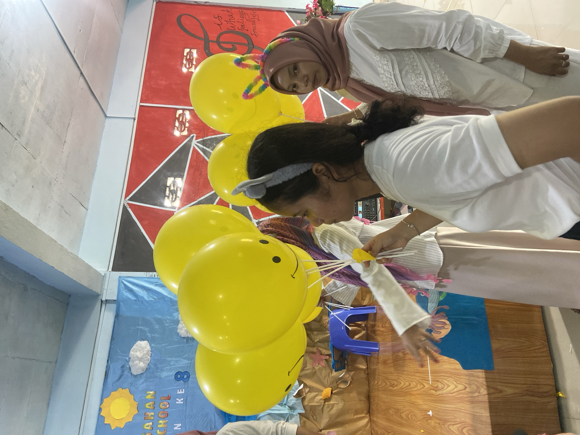 In Tanjung Gading, Indonesia, an event is taking place, possibly a school function, as evidenced by the banner in the background that reads "ASAHAN SCHOOL" and "N KE 8". The scene is decorated with yellow smiley-face balloons, a blue banner depicting a sun and clouds, and a red and black geometric wall art. Three women are visible in the foreground. One woman, in the center, wears a white shirt with vertical pinstripes, a pink scarf, and a gray cat-ear headband. She is holding a yellow balloon string. To her right, another woman is wearing a white shirt with lace detailing and a pink hijab. She has a colorful bunny-ear headband. The third woman, on the far left and only partially visible, wears a white t-shirt and has a gray headband. The overall atmosphere suggests a festive occasion.