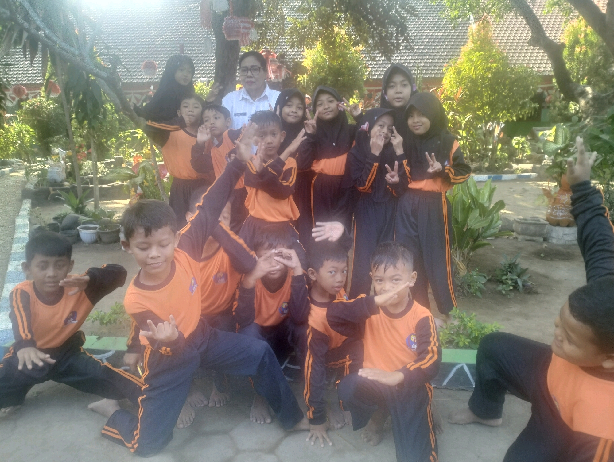 The image depicts a group of Indonesian children, likely students, posing for a photograph outdoors. They are dressed in matching orange and black uniforms, with some girls wearing headscarves. The children are arranged in a semi-circle, some standing and some crouching. Many are making hand gestures. The background suggests a school or community setting, with a building with a tiled roof and lush greenery. The lighting indicates it is daytime, with sunlight filtering through trees. The location is identified as Blabakan, Indonesia. The children appear to be engaged in a playful or ceremonial activity, common in school events.