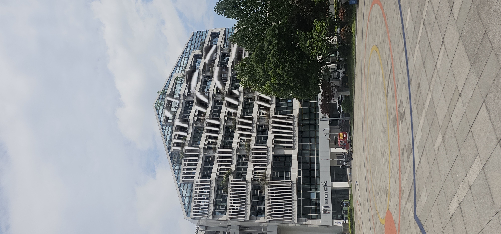 A low-angle, outdoor, full shot depicts a modern, multi-story office building in the Jiulongpo District of Chongqing, China. The building has a stepped, triangular facade with rows of windows covered by horizontal, white slats. The building's base is wider than its top. Some greenery is visible on certain levels. The building's right side features a sign that says "BUICK." A parking lot or paved area with painted lines is in the foreground. A few trees are in the upper right corner, partially obscuring the top of the building. The sky is blue with white clouds.