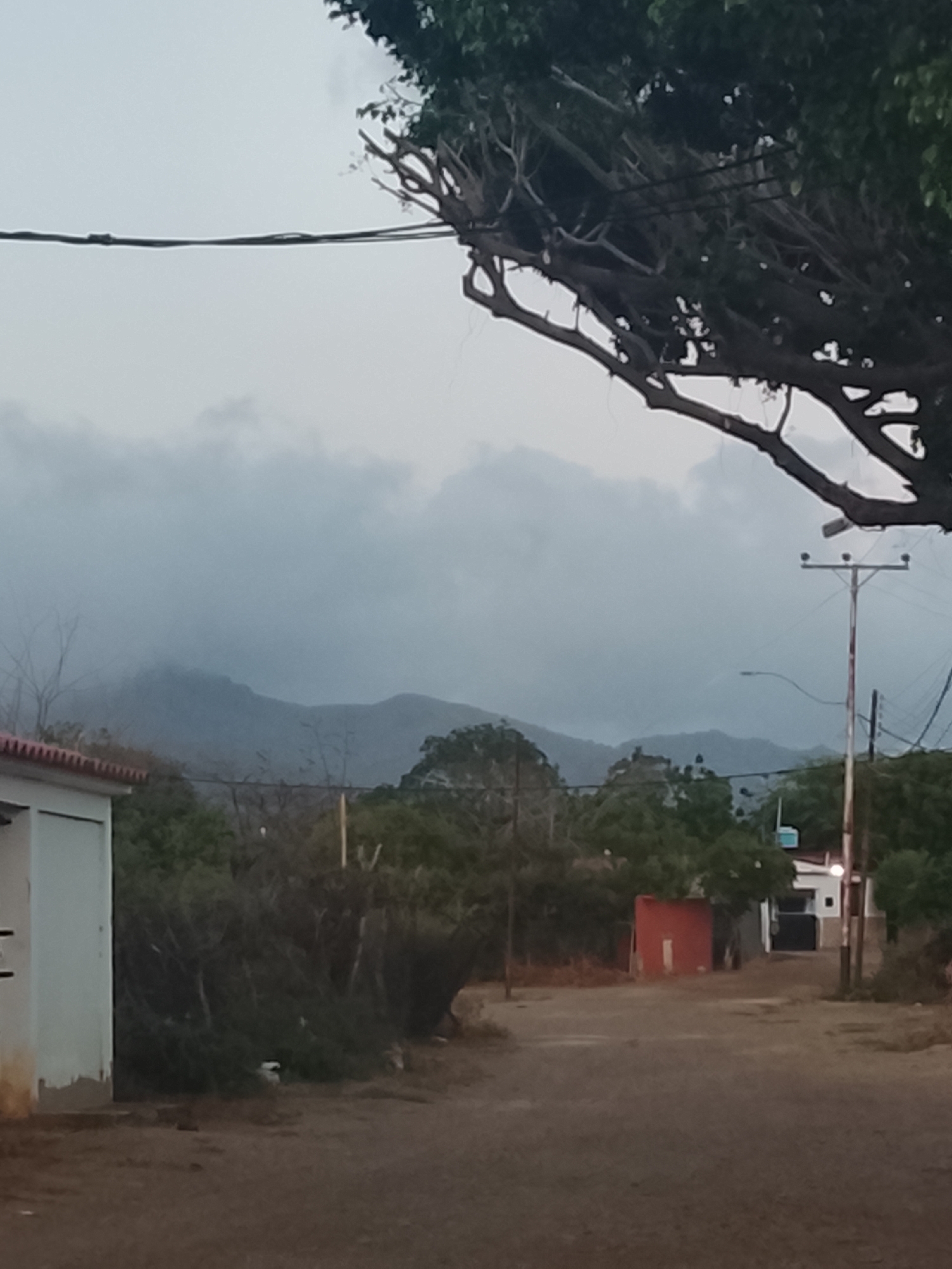 The image shows a street in Altagracia, Venezuela, under a cloudy sky. Mountains are visible in the background, partially obscured by clouds. A tree with thick branches overhangs the upper right portion of the frame. On the left, a white building with a red roof is partially visible. Power lines stretch across the top of the image. Further down the street, a red wall and a white building are present. A utility pole stands on the right side of the road. The road is dirt. There are no people visible.