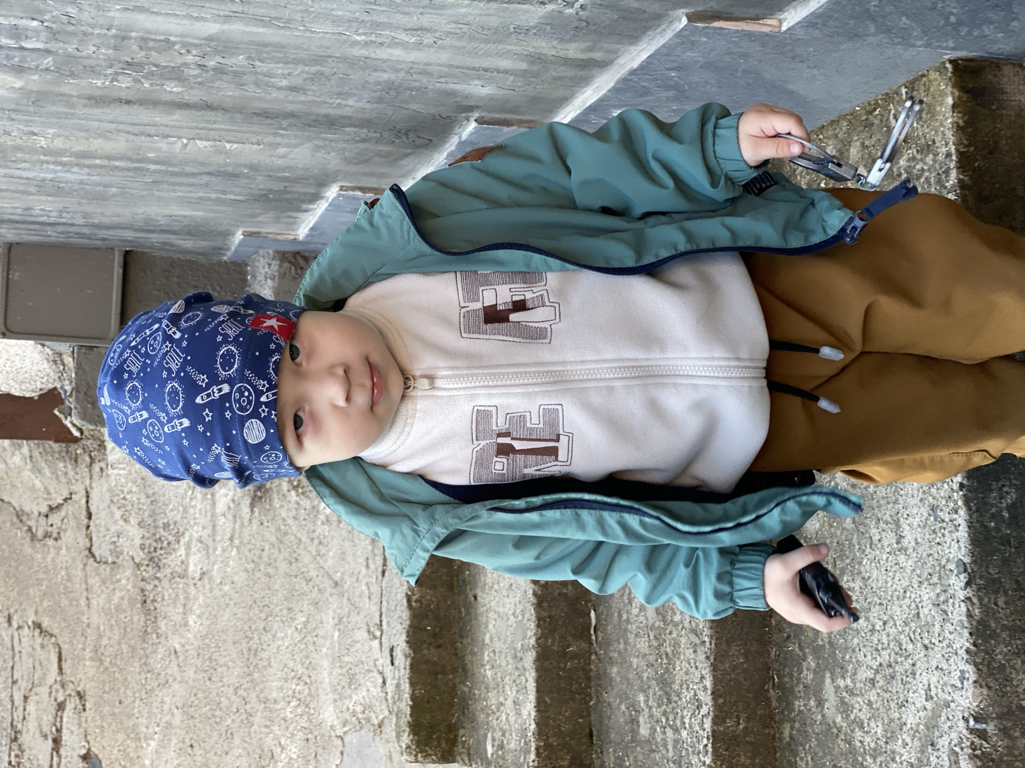 A young child wearing a blue patterned bandana, a white zippered sweatshirt with a brown block design, a teal jacket, and tan pants is sitting on concrete steps. The child is holding a silver manicure tool in their right hand and a black object in their left hand. The steps appear to be outdoors, possibly near a building. The location is specified as Sochi, Russia.