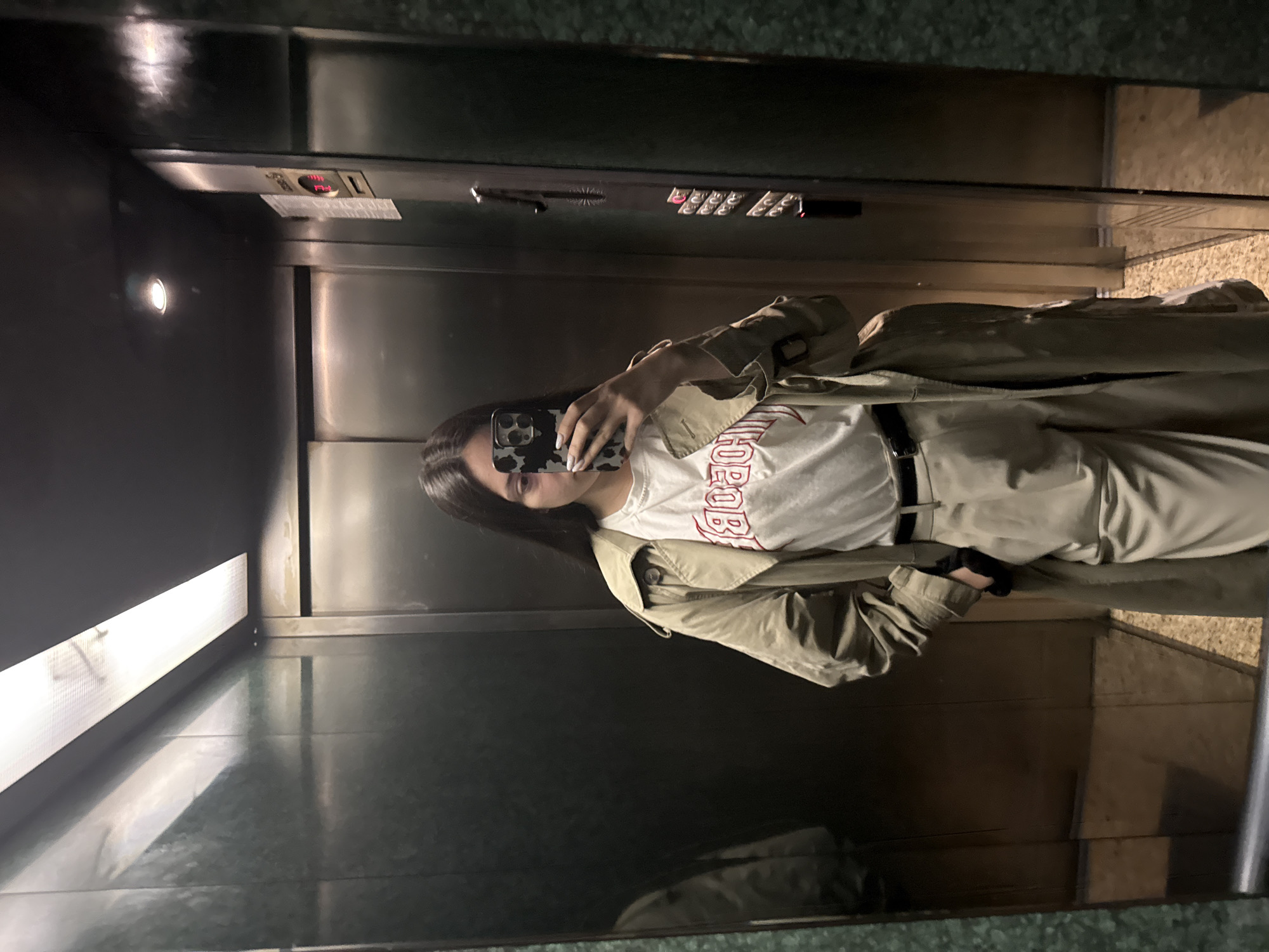 A woman is taking a selfie in an elevator. The elevator has steel walls, a panel with floor numbers, and a light fixture on the ceiling. The woman is wearing a trench coat, a white t-shirt with red lettering, and dark pants with a belt. She is holding a smartphone with a patterned case. The location appears to be inside an elevator in Sofia, Bulgaria.