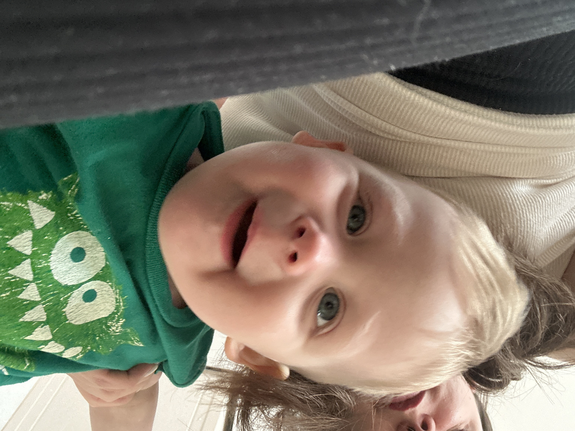 A close-up, eye-level shot captures a happy baby or toddler interacting with an unseen adult, likely a parent. The child, with fair skin, blonde hair, and striking blue eyes, is the central focus, looking directly at the camera with an open-mouthed, joyful expression. They are wearing a vibrant green t-shirt adorned with a large, stylized white monster face design, featuring two prominent circular eyes and triangular shapes.
The image is captured from a perspective looking upwards at the child, suggesting the adult is holding them or lying below them. Partially visible at the bottom and right of the frame is the adult's head, showing dark hair and parts of their chin and mouth, indicating they are looking down at the child. The adult appears to be wearing a light-colored, ribbed garment. Above the child's head, a dark, blurred fabric element occupies the top of the frame.
The setting appears to be indoors, under soft, even lighting, creating an intimate and warm atmosphere. No specific location cues are visible within the image itself, though the location context is Chaikovsky, Russia. The overall emotion conveyed is one of delight and affectionate connection. There is no visible text.