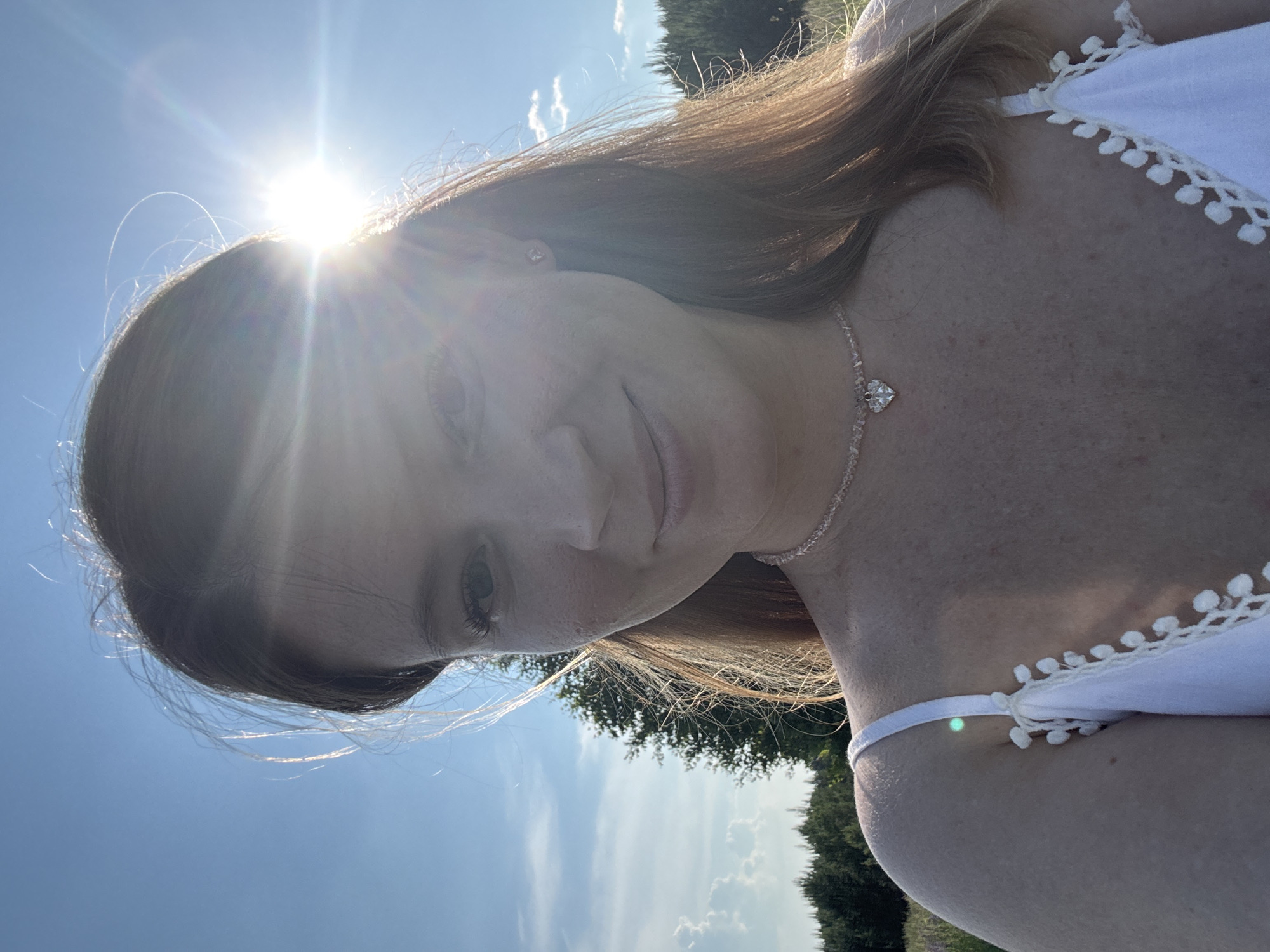 A woman, appearing to be in her early twenties, is captured in a close-up selfie taken outdoors. She has light skin, freckles, and shoulder-length auburn hair. She is wearing a white sundress with a delicate lace trim and a thin pink beaded necklace with a heart-shaped pendant. The sun is bright and low in the sky, partially obscuring her face and creating lens flare. Behind her, a blue sky with scattered white clouds is visible, along with a treeline and a single tall tree to her left. The background suggests a rural or natural setting, consistent with the village of Van'ki, Russia, which is known for its picturesque landscapes. The lighting and pose indicate a casual, sunlit moment.