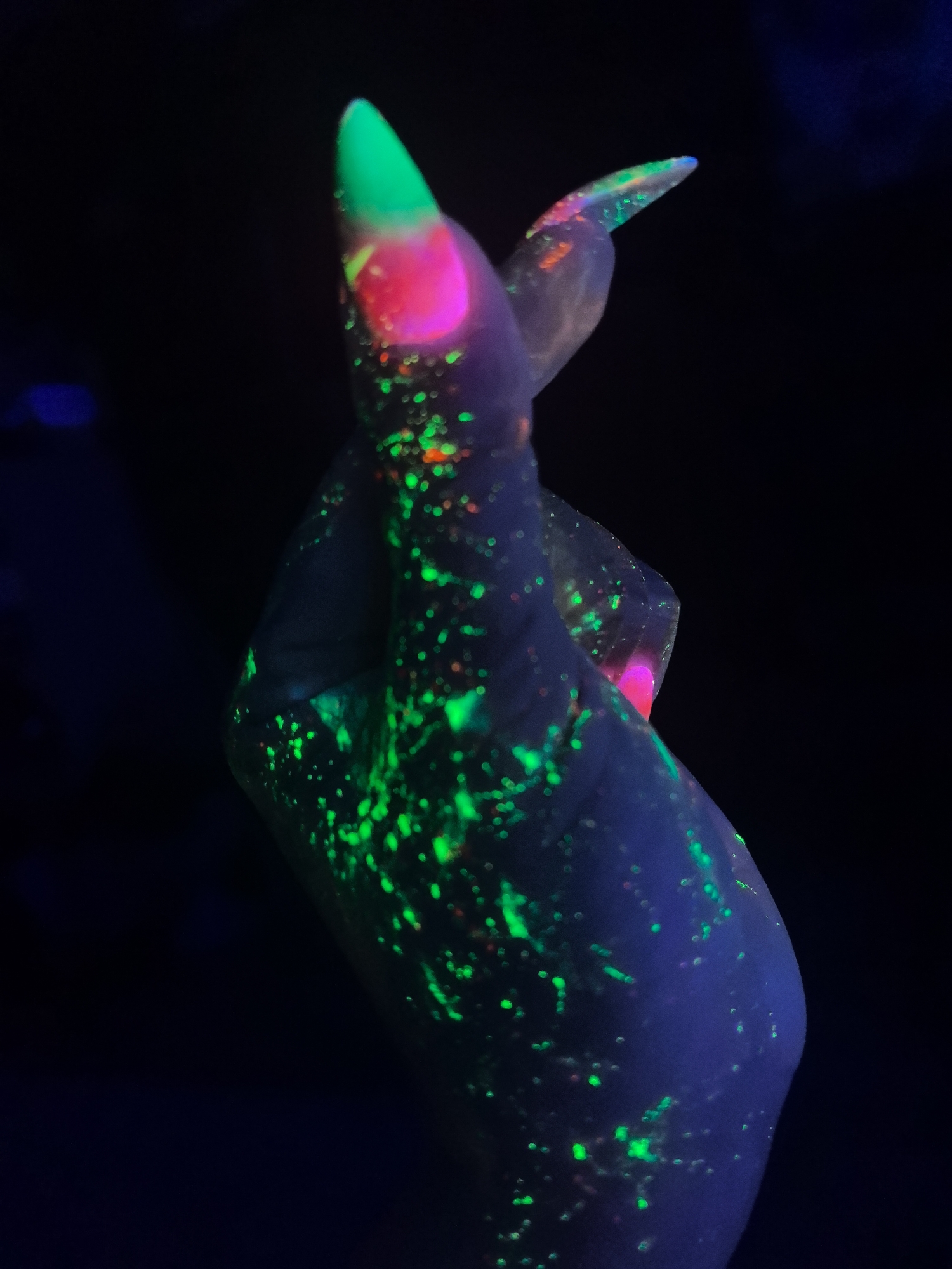 The image shows a human hand in a dark environment, likely under ultraviolet (UV) light. The hand and fingernails are decorated with fluorescent paint in shades of green, pink, and orange, which glow brightly under the UV light. The background is black, suggesting a darkened room or nighttime setting. The hand is positioned with two fingers crossed. There is no location information, so I cannot specify if the location is in Noviy, Russia.