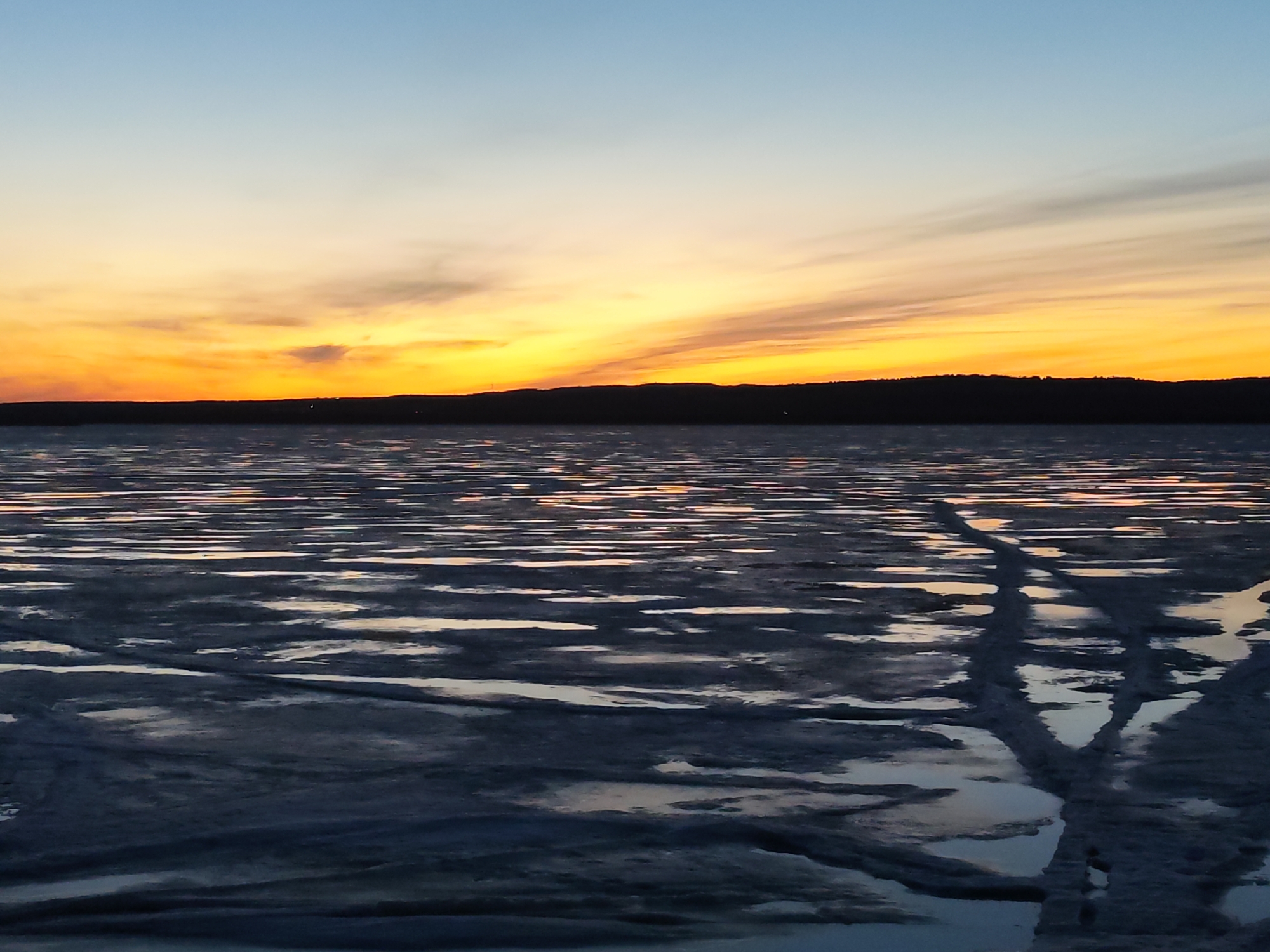 The image shows a body of water, likely a lake or reservoir, partially covered in ice or slush. The water reflects the colors of the sunset, which dominates the upper portion of the image. The sky transitions from light blue at the top to a gradient of orange and yellow closer to the horizon. A dark, silhouetted landmass is visible on the horizon, separating the sky from the water. The foreground features textured ice and snow, with visible tracks or patterns indicating movement across the surface.
Without more information, I cannot place the location as Chaikovsky, Russia, but the scene suggests a cold climate region.