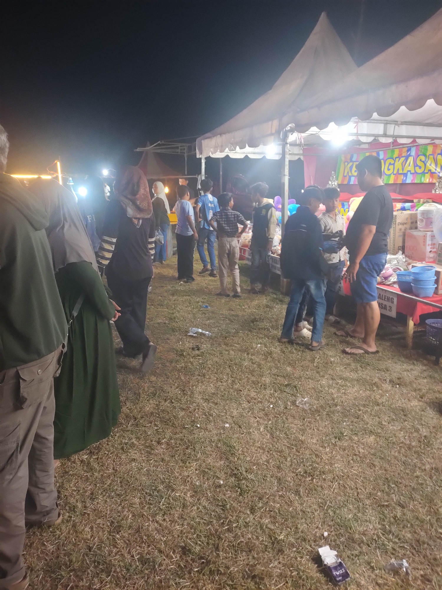 The image captures a nighttime scene at what appears to be an outdoor market or event in Subang, Indonesia. Several tents are set up, illuminated by artificial lighting, casting shadows on the dry, mowed grass covering the ground. A banner with colorful lettering, partially obscured, is visible on one of the tents.
A diverse group of people, including adults and children, are present. Some individuals are walking, while others appear to be congregating near stalls or tents. Clothing varies, with some people wearing headscarves and others in casual attire. The subjects are mostly facing away from the camera or are partially visible, making detailed identification difficult.
In the foreground, a person in a green hoodie and khaki pants is standing. Nearby, a person in a long, dark green dress is also visible. The ground is scattered with some litter, including a crumpled paper and a discarded cigarette pack.
The overall impression is of a busy, casual evening gathering in an open area, likely a local community event or a night market. The lighting and the nature of the structures suggest a temporary setup.