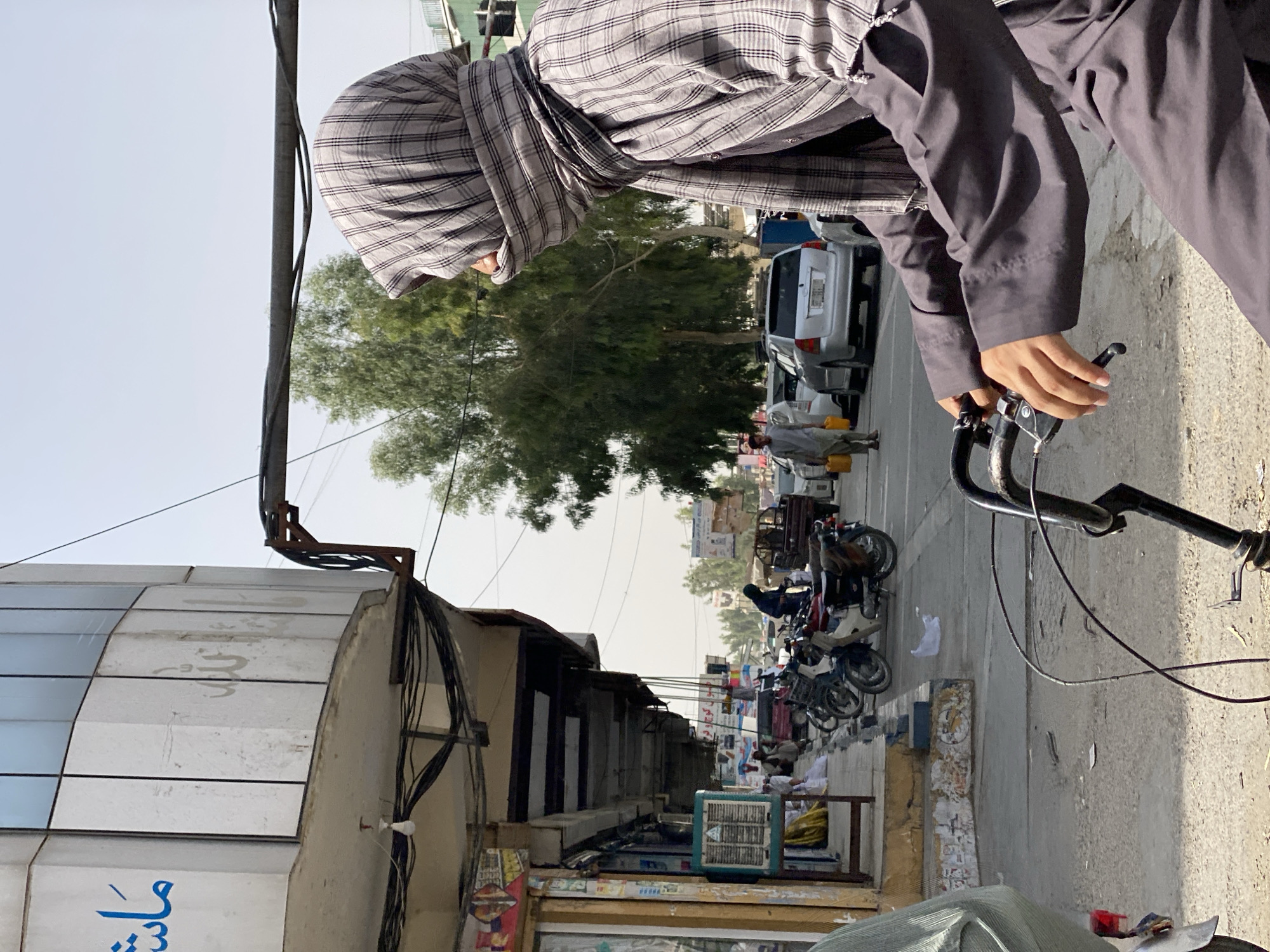 The image shows a street scene in Kandahar, Afghanistan, on a sunny day. In the foreground, a person wearing a gray and white plaid scarf over their head and face, and a dark gray tunic, rides a bicycle. The person's hands are on the handlebars, and the bicycle's frame and part of the rear wheel are visible.
The background depicts a street with several parked motorcycles and cars. To the left, a building with white tiled facade and blue Arabic script is visible. Several shops with awnings line the street. People are walking on the sidewalk, and some are gathered near the shops. A man in a light-colored tunic and carrying a yellow jerrycan walks on the sidewalk to the left of the parked cars. Trees line the street in the distance, and overhead power lines are visible. The overall scene suggests a typical day in a bustling urban environment.