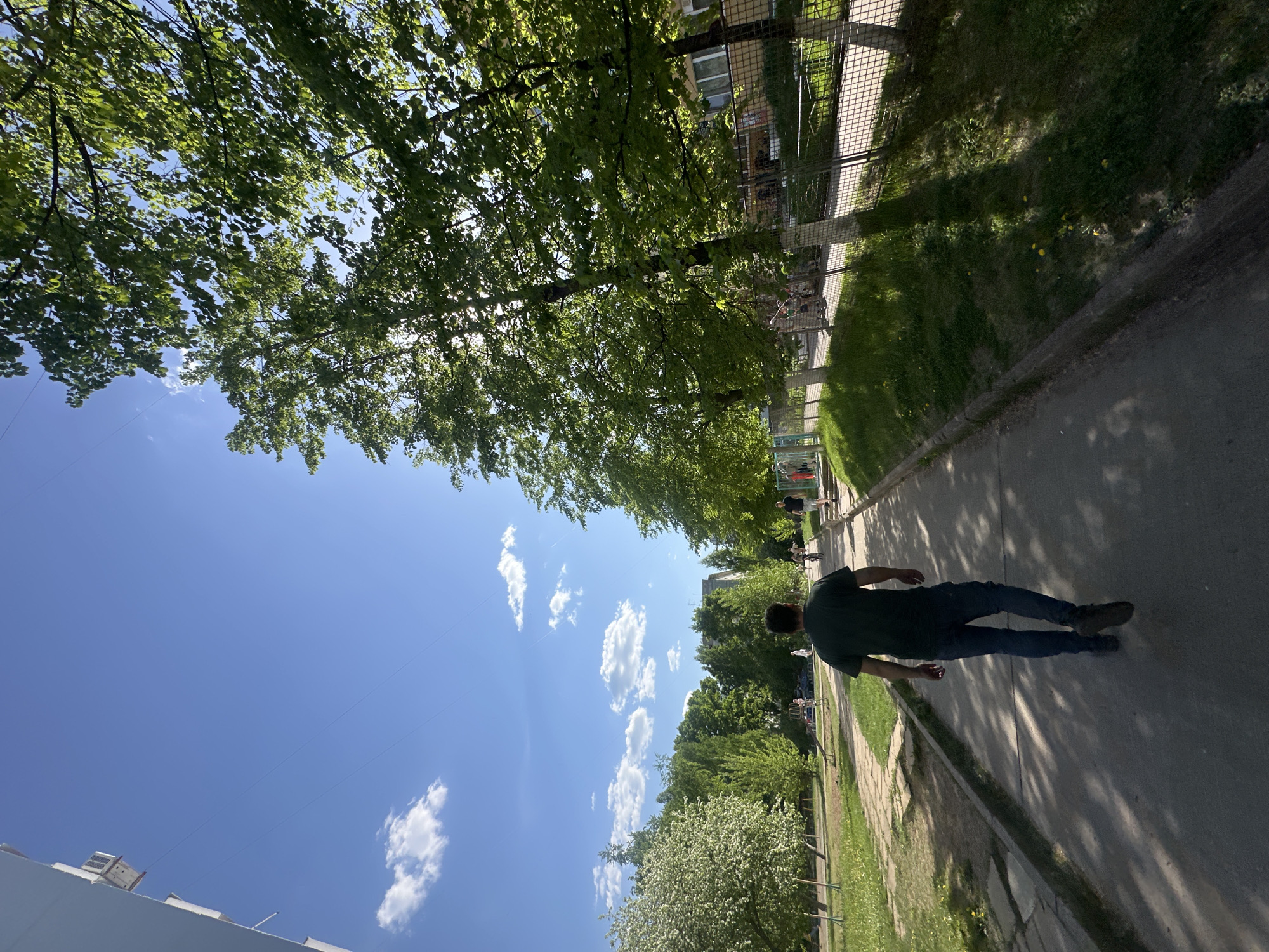 The image shows a street scene in Chaikovsky, Russia, taken on a sunny day with a blue sky dotted with scattered cumulus clouds. The viewpoint is angled upwards and to the left. A man, seen from the back, is walking along a paved pathway in dark clothing, including a short-sleeved shirt and jeans. To the right of the pathway is a grassy area bordered by a low concrete wall. Beyond the grass, there is a chain-link fence, trees, and what appears to be the side of a building. To the left of the pathway are trees with dense foliage that filters the sunlight. The foreground also contains the edge of a building. In the distance, other people can be seen on the pathway.
