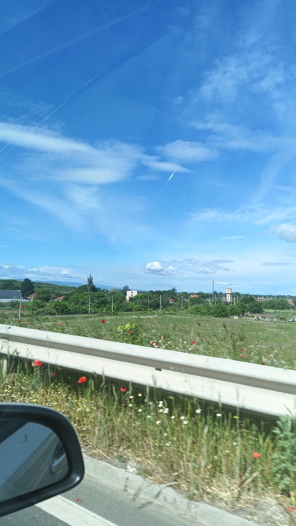 View from a vehicle passing through the outskirts of Popovitsa, Bulgaria. The foreground shows a section of a guardrail along a road, beyond which is a grassy area with red poppy flowers and white wildflowers. In the midground, there are clusters of trees and vegetation, interspersed with occasional buildings. In the background is a clear blue sky with sparse white clouds and contrails. A car's side mirror is visible in the lower left corner of the frame.