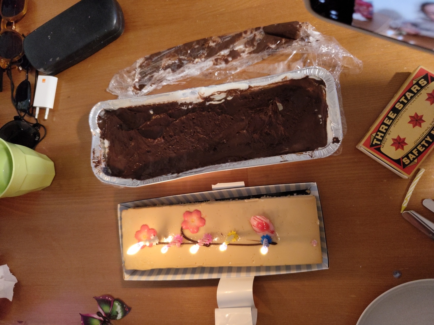 The image presents an overhead view of a wooden table, suggesting an indoor setting for a celebration or gathering.
At the center, two cakes are visible. One is a large, rectangular chocolate dessert served in a silver aluminum foil pan. It features a rich, dark brown top layer, possibly frosting or fudge, with a lighter, creamy layer visible beneath. The other cake, placed in a striped cardboard box, is also rectangular and appears lighter in color, possibly a cheesecake or a pale-frosted sponge cake. This cake is decorated with several small, colorful candles, some burning, alongside delicate pink floral and green leaf-like embellishments.
Surrounding the cakes are various personal items. To the left, there's a black hard case for sunglasses and a pair of dark sunglasses, a white USB wall charger, and a light green plastic cup. A small piece of crumpled white paper lies near the cup. In the top right corner, a partial view of a smartphone shows a reflection of a person's face. To the right of the chocolate dessert, a vintage-style box of "THREE STARS SAFETY MATCHES" is visible, bearing the text "MADE BY JONKÖPING, SWEDEN - VULCAN GS". A partially burnt yellow and white striped candle rests next to the matchbox. Further right, parts of a gray plate and a metal serving utensil are seen. A decorative butterfly-shaped object with purple and green wings is situated in the bottom left corner.
The presence of lit candles on a cake, along with multiple desserts, strongly indicates a celebratory event, most likely a birthday party or a festive gathering. The atmosphere appears casual and joyful. The lighting, with active candles, suggests it could be evening or a dimly lit indoor space.