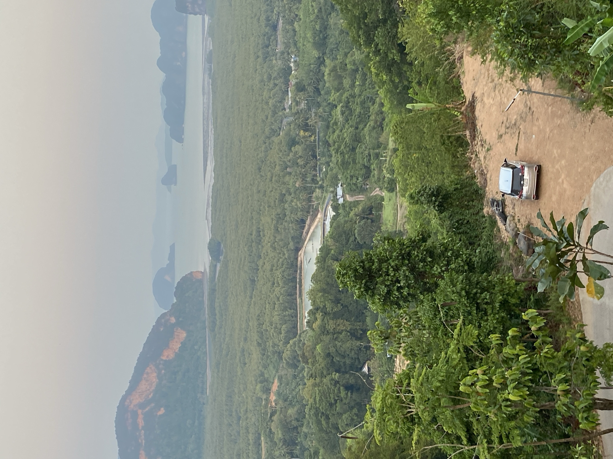 The image depicts a landscape in Khlong Khian, Thailand, viewed from an elevated position. In the distance, there is a body of water, possibly a bay, with islands or landmasses visible through a hazy atmosphere. The middle ground is dominated by dense green vegetation, likely a forest or mangrove area. In the foreground, a sandy or dirt path leads to a parked silver vehicle. Trees and shrubs line the side of the path. The overall impression is of a natural environment with minimal signs of human activity apart from the road and the car.