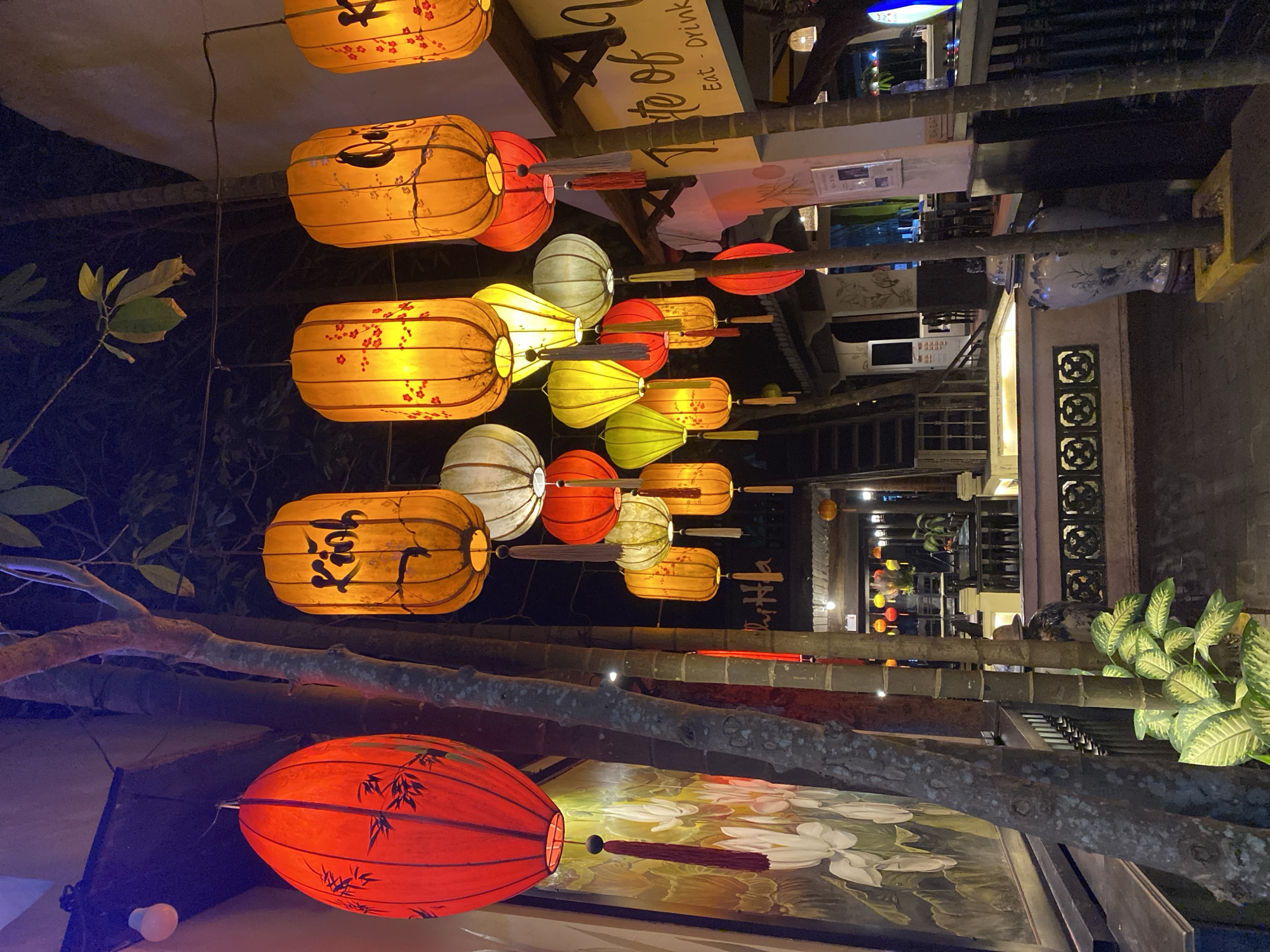 An exterior shot of a restaurant in Da Nang, Vietnam, at night, showcases a vibrant scene illuminated by numerous hanging lanterns. A large red lantern with black patterns of bamboo is prominently displayed on the left. To its right, several cylindrical lanterns in shades of orange and yellow are strung together, some adorned with delicate red floral designs and others with black calligraphy. White and green lanterns are also visible amidst the colorful array. The background reveals the restaurant's interior, with tables and chairs set up and further decorative lighting. A large mural featuring white lotuses is painted on the wall to the left of the red lantern. The overall ambiance is festive and inviting, characteristic of a traditional Vietnamese night market or restaurant setting.