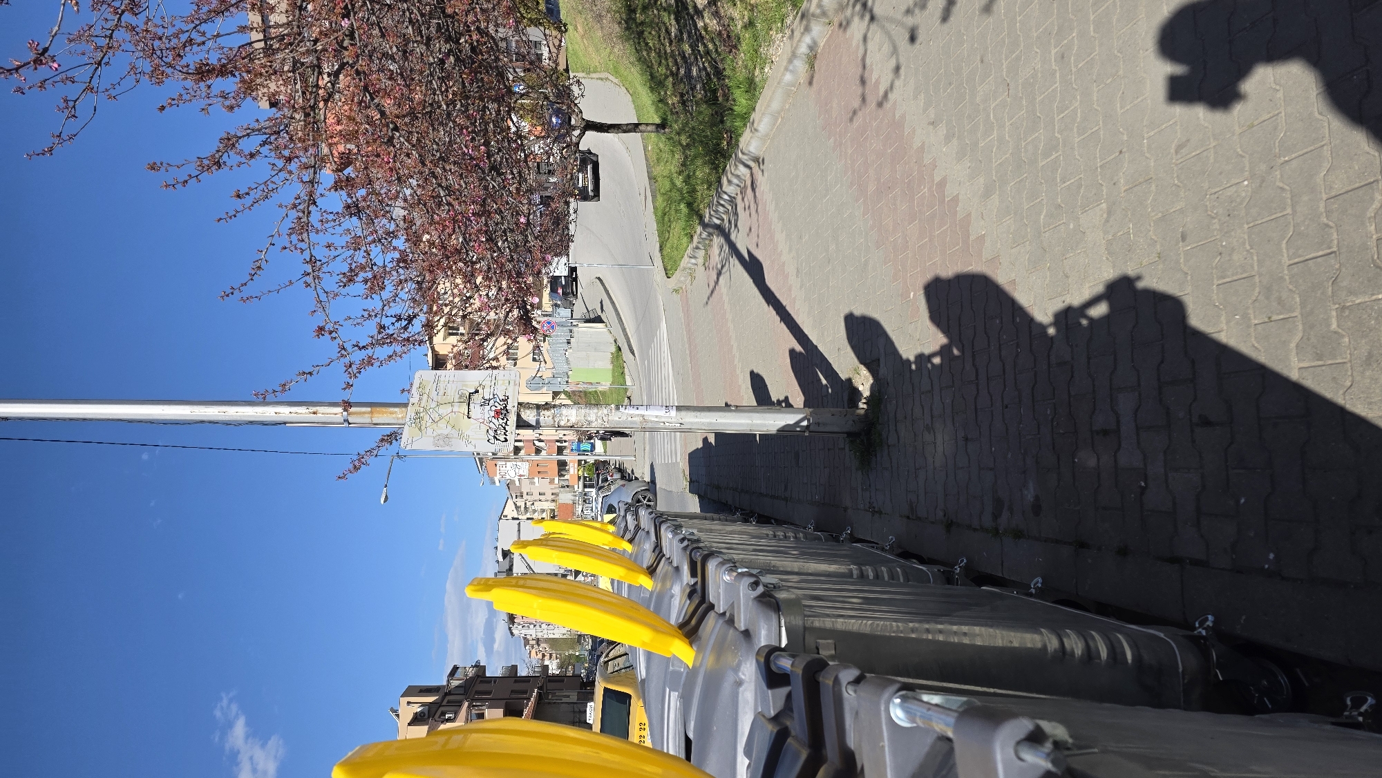 The image shows a row of gray and yellow waste bins on the sidewalk of a street in Blagoevgrad, Bulgaria. The street has a paved sidewalk and a road with cars parked on either side. A utility pole stands along the sidewalk, and a blooming tree overhangs the scene. The sky is blue with scattered clouds. The shadow of the photographer or photographer's device and person taking the photo is visible on the sidewalk. Buildings line the street.