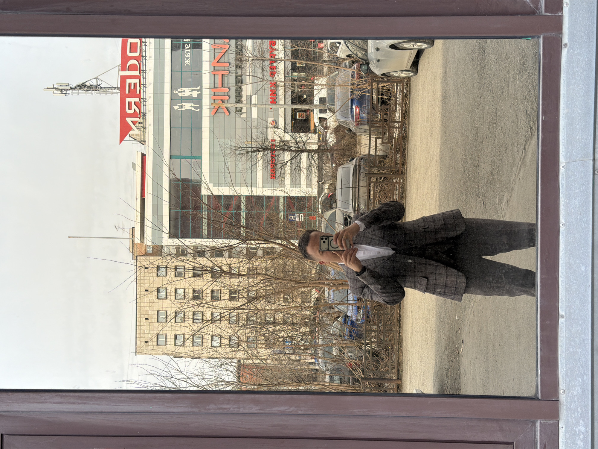 A man in a checkered suit takes a selfie in a window. In the background, a multi-story building with a large sign that reads "О’КЕЙ" (O'KEY) is visible. Several cars are parked on the street. The presence of the "O'KEY" hypermarket suggests the city of Perm, Russia, as it is a common retail chain in that country. A neighboring building with multiple windows can also be seen. Bare trees are visible, suggesting the photo was taken during late autumn or early spring.