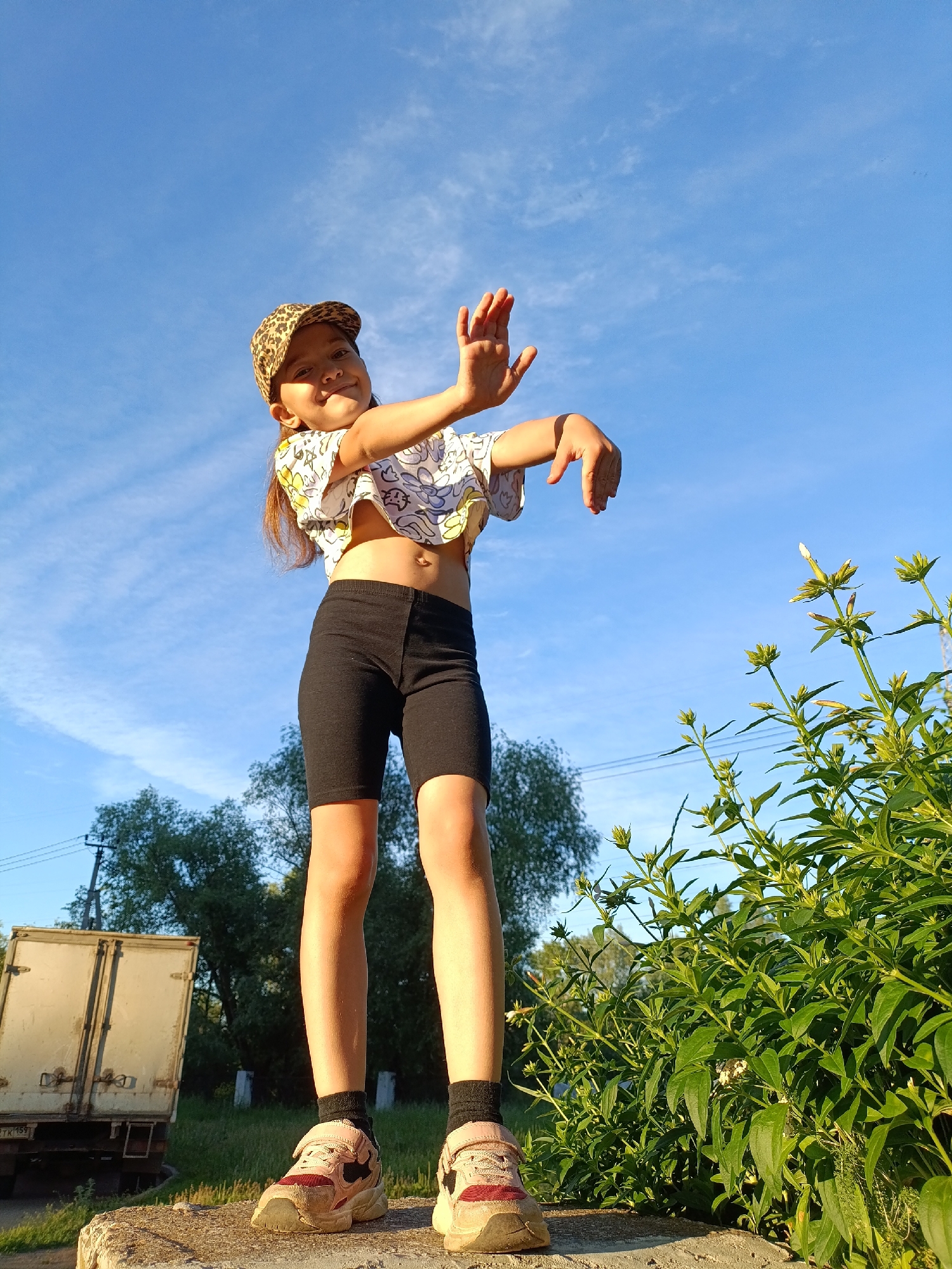 A young girl, wearing a leopard print baseball cap, a cropped t-shirt, black shorts, and pink sneakers, stands with her arms raised and hands open, looking towards the camera. She is positioned on a concrete surface with a lush green bush to her right and a white truck in the background. The scene is set against a bright blue sky with wispy clouds, suggesting it is daytime and outdoors. The location is identified as being in Chaikovsky, Russia.