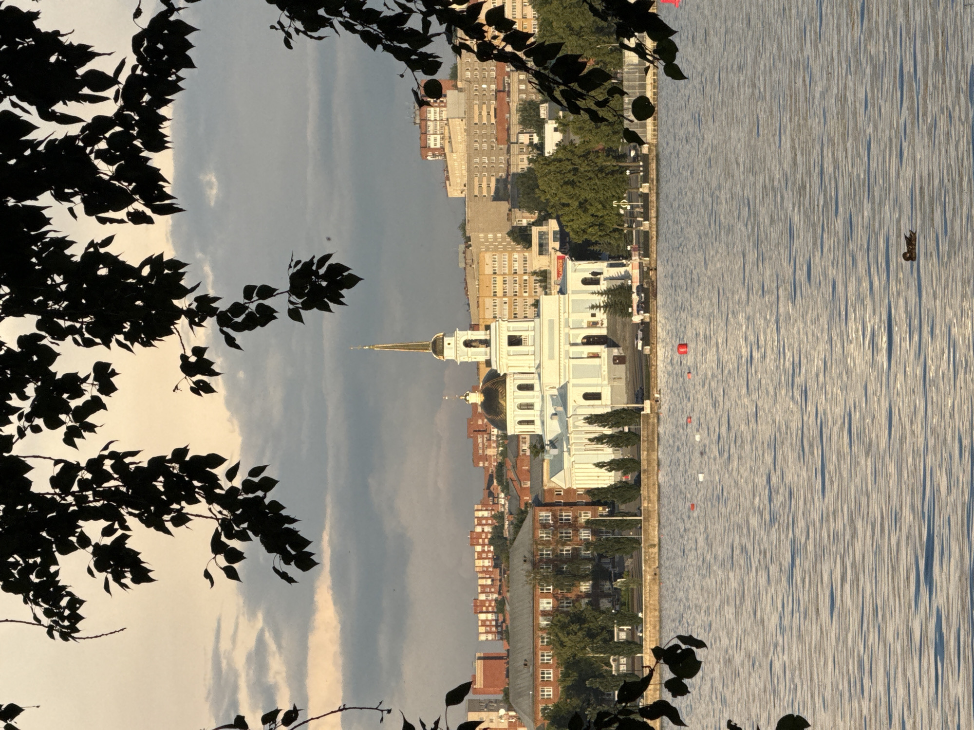 In Votkinsk, Russia, a serene lake reflects the sky and surrounding cityscape. In the foreground, dark tree branches with leaves frame the scene, casting shadows. The lake is rippled, with a lone duck swimming in the lower right portion of the image. Across the water, a white church with a golden dome and steeple stands prominently, situated along the shore. Behind the church, a dense array of city buildings, varying in height and architectural style, stretches across the horizon. The sky above is a mixture of soft, muted clouds, with hints of golden light suggesting either sunrise or sunset. A stone embankment separates the water from the urban area, lined with trees and a paved walkway. Several buoys, red and white, are visible in the water, marking an area.