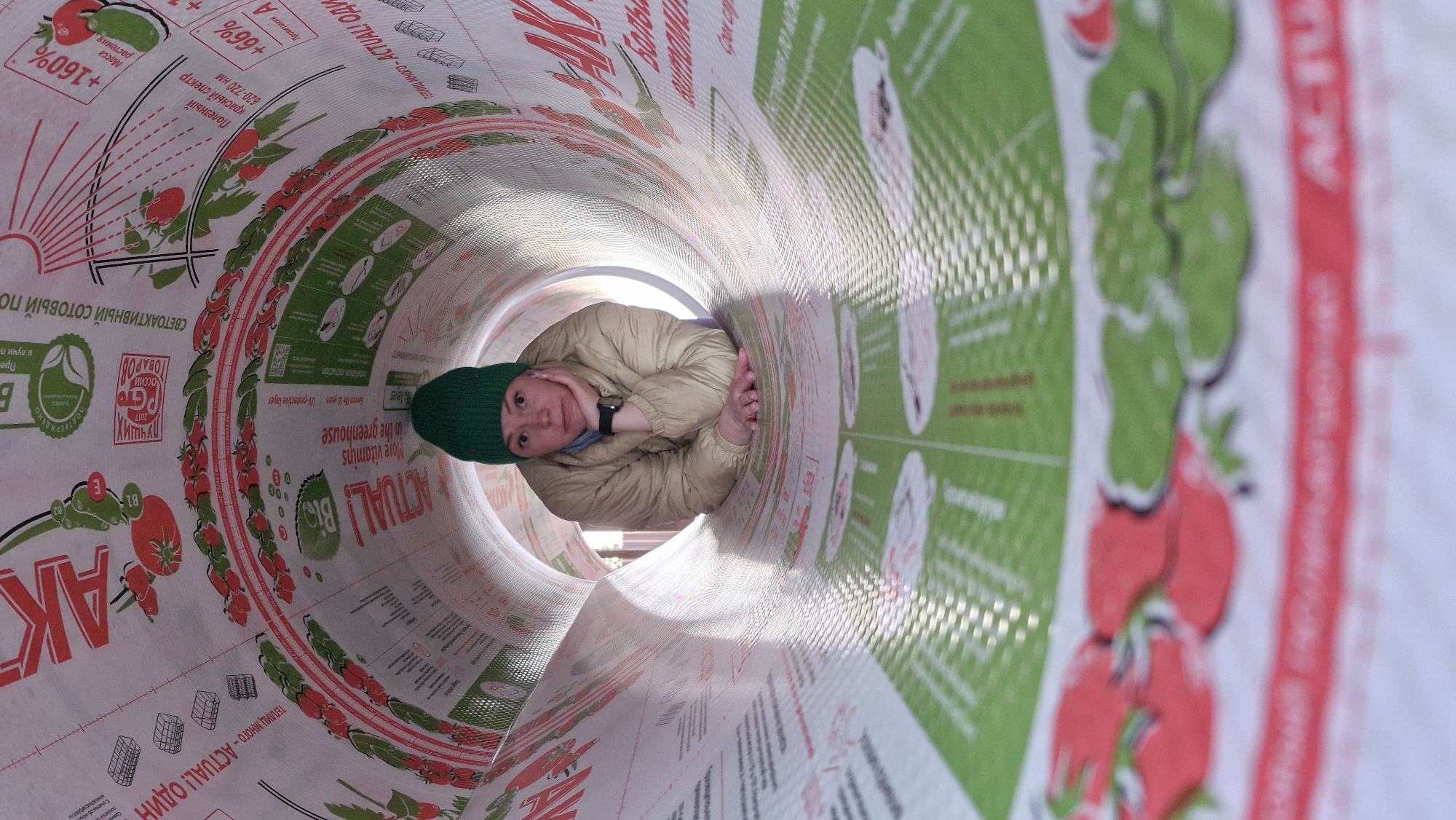 The image shows a person lying inside a large cylindrical tube. The person, likely female, wears a green knit hat and a beige jacket. Their head rests on one hand and a wristwatch is visible on the other wrist. The inside of the tube is printed with text and graphics, including images of tomatoes, text in Russian, and the word "ACTUAL!" in English. The background surrounding the opening of the tube appears to be outside, possibly a park or garden area. The OCR text identifies Russian words such as "ТЕПЛИЦ МНОГО - ACTUAL! ОДИН" which translates to "Many greenhouses - ACTUAL! One". The location is identified as Otchor, Russia.