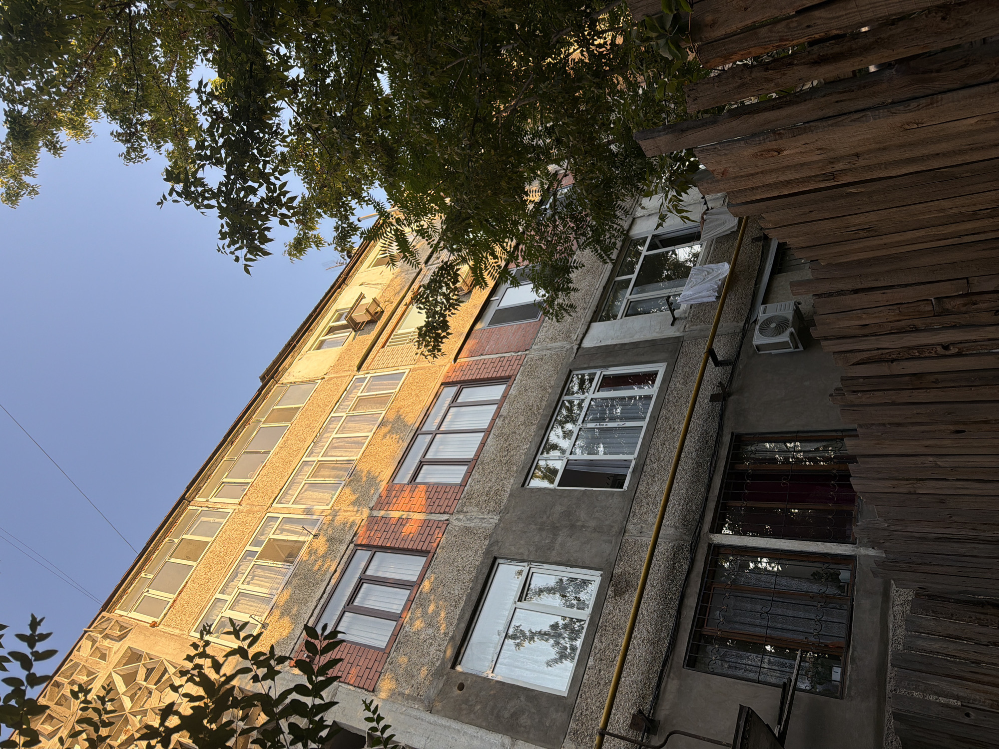 This is a low-angle shot of a multi-story apartment building in Shymkent, Kazakhstan. The building's facade is a mix of rough concrete and brickwork, with several windows and balconies visible. The upper floors are bathed in the warm light of the setting sun, creating a stark contrast with the shadowed lower levels. A large tree with dense green foliage partially obscures the right side of the building. A weathered wooden fence runs along the bottom of the frame, separating the viewer from the building. Laundry is hung on a line outside one of the windows. An air conditioning unit is mounted on the wall below another window. The sky is clear and blue.