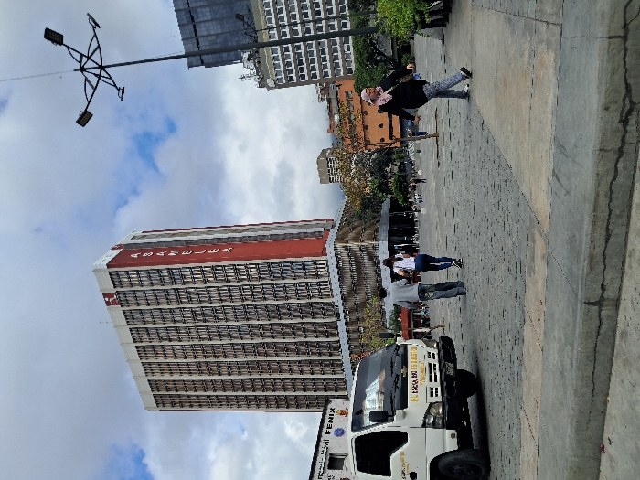 The image presents an urban street scene in Caracas, Venezuela, centered on the area adjacent to the Federal Legislative Palace. A tall, multi-story skyscraper, the National Assembly building, dominates the background, featuring a facade of numerous windows and a prominent vertical red stripe on its right side with "ASAMBLEA" written in white capital letters. The sky above is partly cloudy, with white clouds against a blue backdrop.
In the mid-ground and foreground, a paved plaza or street area is visible, with a concrete curb showing minor cracks running along the bottom. On the left, a white light-duty truck, appearing to be a delivery or utility vehicle, is positioned. Text such as "EL ESCUDO ES NUESTRO" is partially visible on its front.
Three individuals are prominent in the mid-ground: two figures, a male in a grey hoodie and jeans and a female in a white top and dark pants, walk away from the camera's perspective. To their right, a third individual, a female wearing a black top, patterned light pants, and a light-colored head covering, walks towards the right side of the frame. Multiple indistinct pedestrians are visible further in the background within the plaza. A tall, black streetlight with four radiating fixtures stands prominently in the upper right quadrant of the image. Other urban buildings are discernible in the background, beyond the main plaza area.