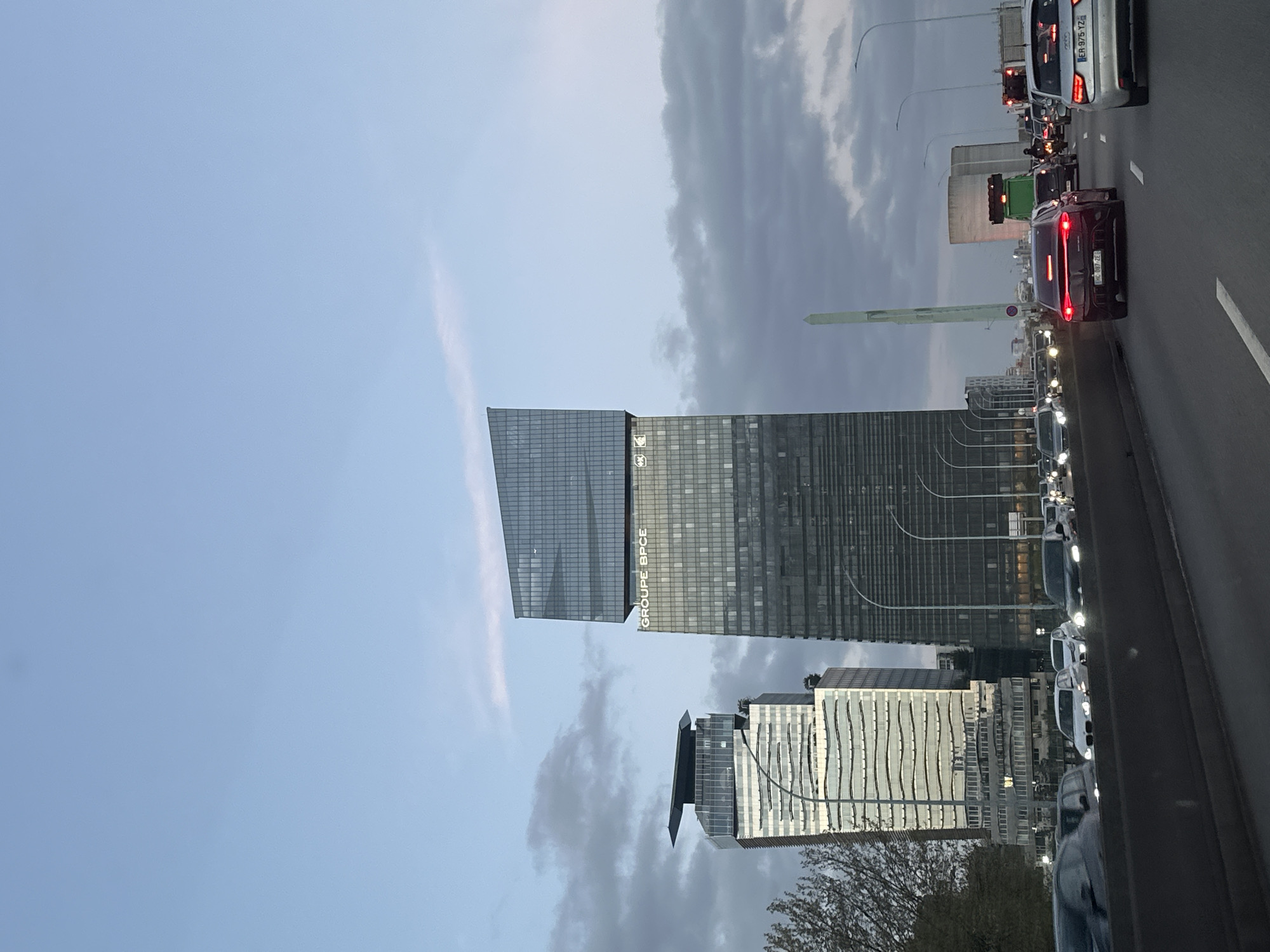 The image shows a view from a vehicle on a highway in Paris, France, facing the city skyline. Traffic is congested. Prominent features include the modern architecture of high-rise buildings, notably the Group BPCE tower. The sky is overcast with clouds. In the foreground, cars are lined up on the highway, with their tail lights visible. A tall, light-colored monument is also visible in the distance.
