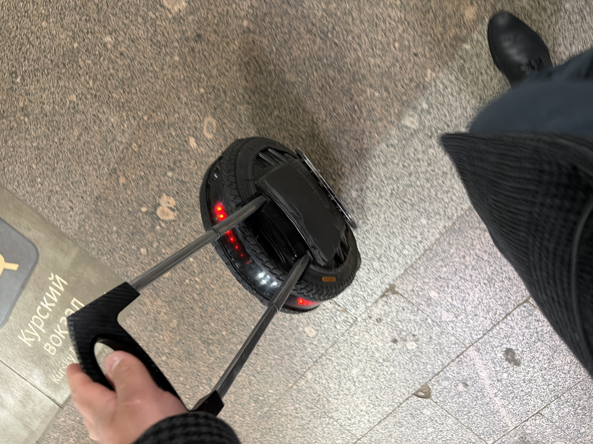 A person is standing on a speckled gray floor next to a black, single-wheeled, self-balancing scooter. The scooter is fitted with an extendable handle, which the person is holding with their left hand. The scooter has red lights around the edge of the wheel. The person is wearing a black boot on their right foot and black fabric is visible next to their leg. A sign partially visible on the floor to the left reads "Курский вокзал", which translates to "Kursky Station".