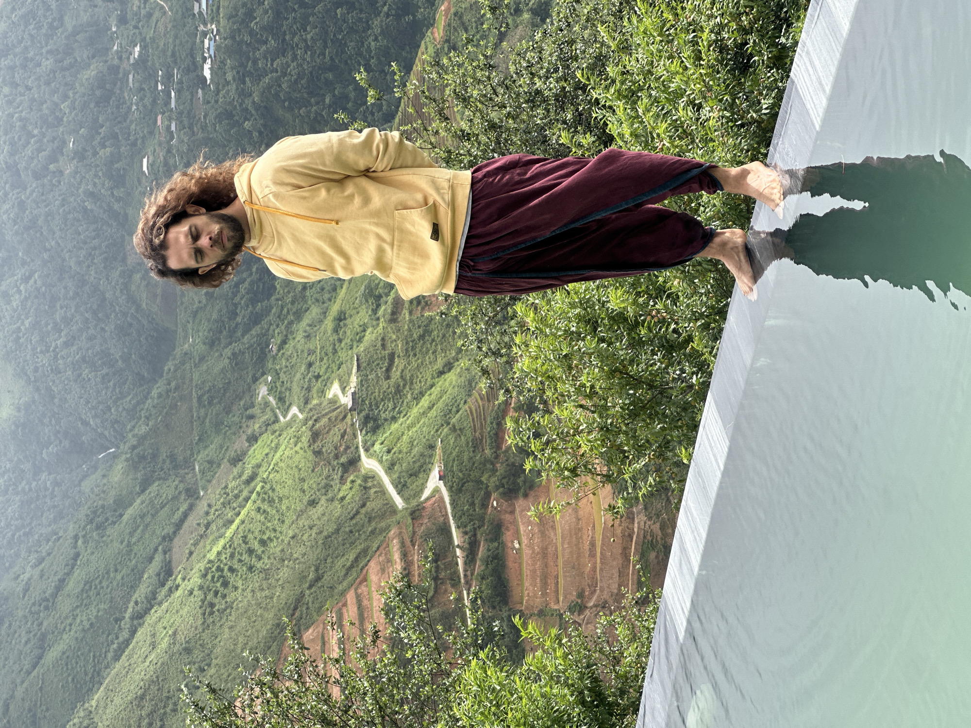 A man with curly brown hair and a beard stands on the edge of an infinity pool, looking down with his eyes closed. He is wearing a yellow hooded sweatshirt and dark red loose-fitting pants. The pool's water reflects his image. Behind him, the landscape of Ta Xua, Vietnam, unfolds with lush green mountains, terraced fields, and a winding road. The sky is overcast. The man's feet are bare and touching the water's edge.