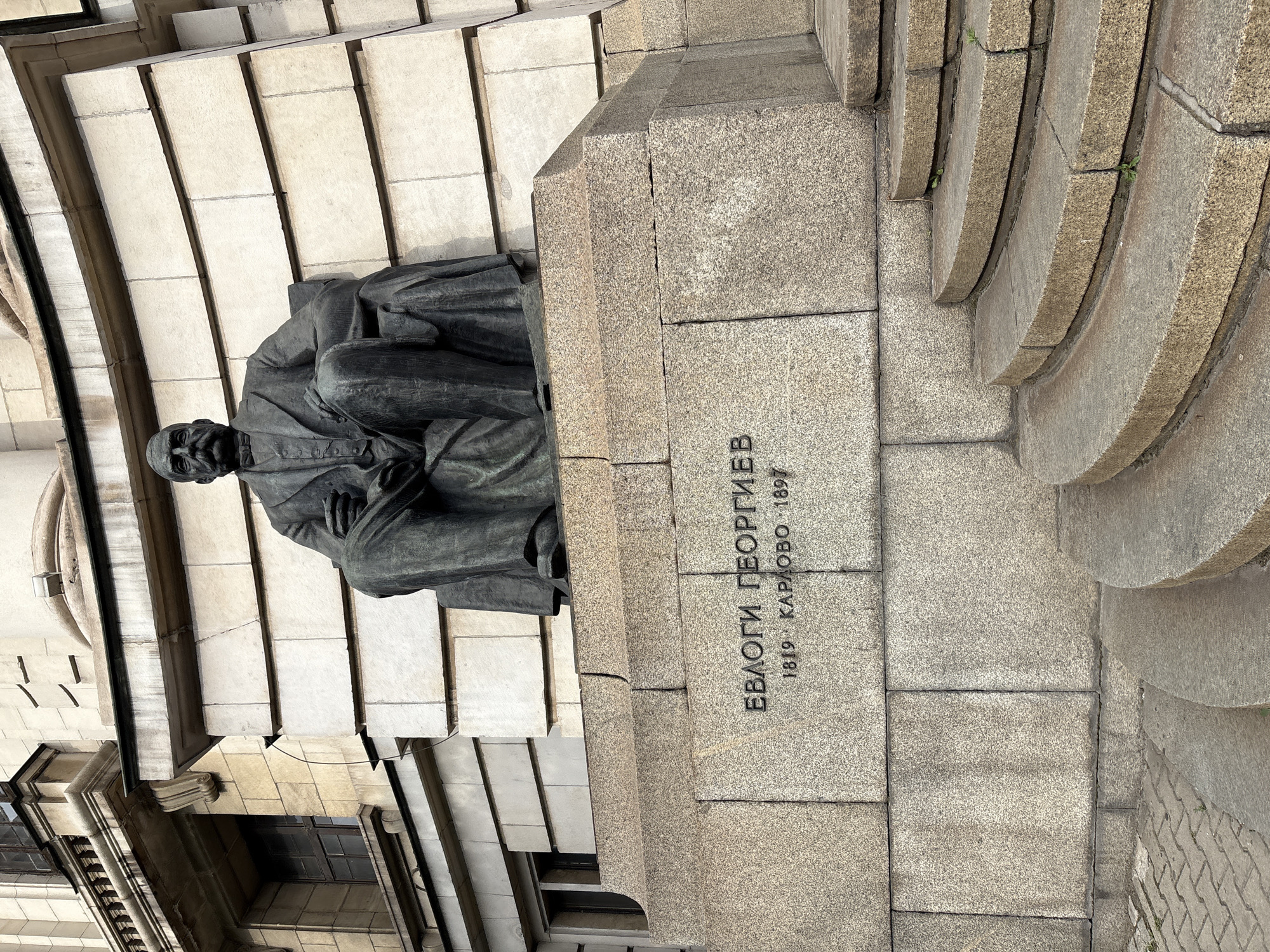 The image shows a bronze statue of a man seated on a stone pedestal in front of a building. The man is wearing a suit and has a mustache. The pedestal has an inscription that reads "ЕВЛОГИ ГЕОРГИЕВ 1819 - КАРЛОВО - 1897". The building behind the statue is made of stone blocks and has windows with decorative elements. There are also stone stairs leading up to the pedestal. This is the statue of Evlogi Georgiev located in front of the Sofia University in Sofia, Bulgaria.