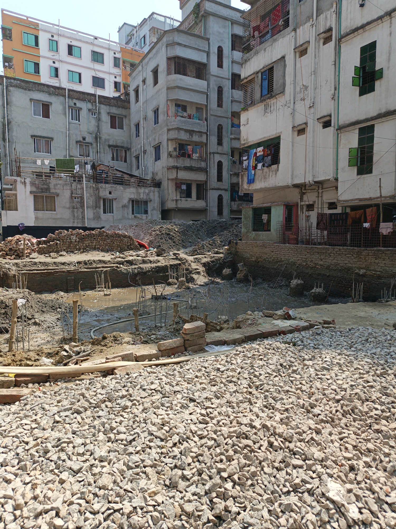 The image shows a construction site in Dhaka, Bangladesh. The foreground is covered with gravel. In the middle ground, there is exposed soil, rebar, and bricks, suggesting the beginnings of a foundation. Water has pooled in the excavated area. In the background are densely packed, multi-story residential buildings with visible signs of aging and disrepair. Laundry is hanging from windows and balconies. One person is visible in the distance near a pile of bricks.