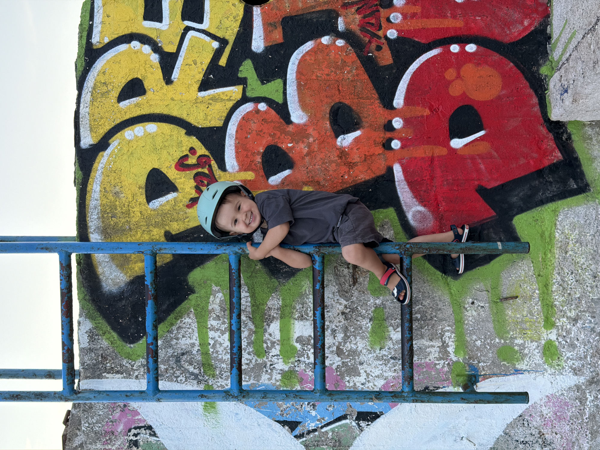 A joyful young child, wearing a light blue helmet, a dark gray t-shirt, and dark shorts, is playfully lying on a blue metal ladder. The child beams at the camera with a wide, happy smile, resting his head on his left arm. He is wearing dark blue sandals with red accents.
The ladder, which shows significant wear with peeling blue paint and rust, is set against a large, textured concrete or stone wall. This wall is extensively covered in colorful graffiti art, featuring large, stylized block letters and abstract shapes. Predominant colors in the graffiti include bright yellow, orange, red, black, white, and green. The art appears to be layered, suggesting multiple applications over time. The background on the left side reveals a bright, clear sky, indicating daytime and fair weather. The scene exudes a sense of carefree playfulness in an urban or public setting in Budva, Montenegro. No specific legible words are clearly discernible within the graffiti, only abstract letterforms.